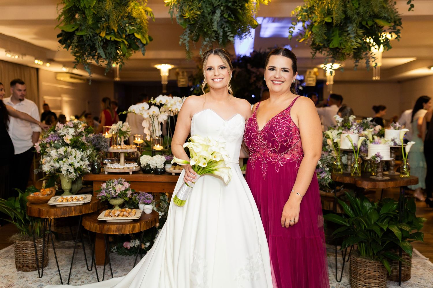 Bride in white gown holding bouquet poses with woman in red dress at elegant wedding reception.