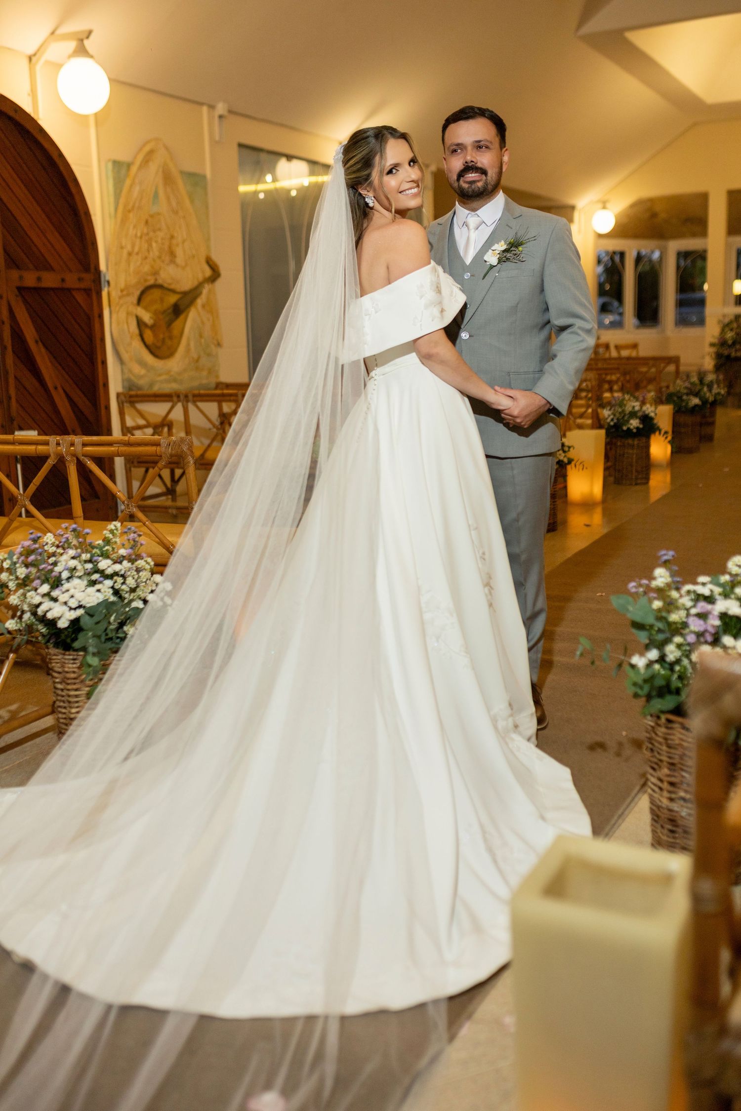 Bride in white off-shoulder gown with long veil stands with groom in gray suit at elegant wedding venue.