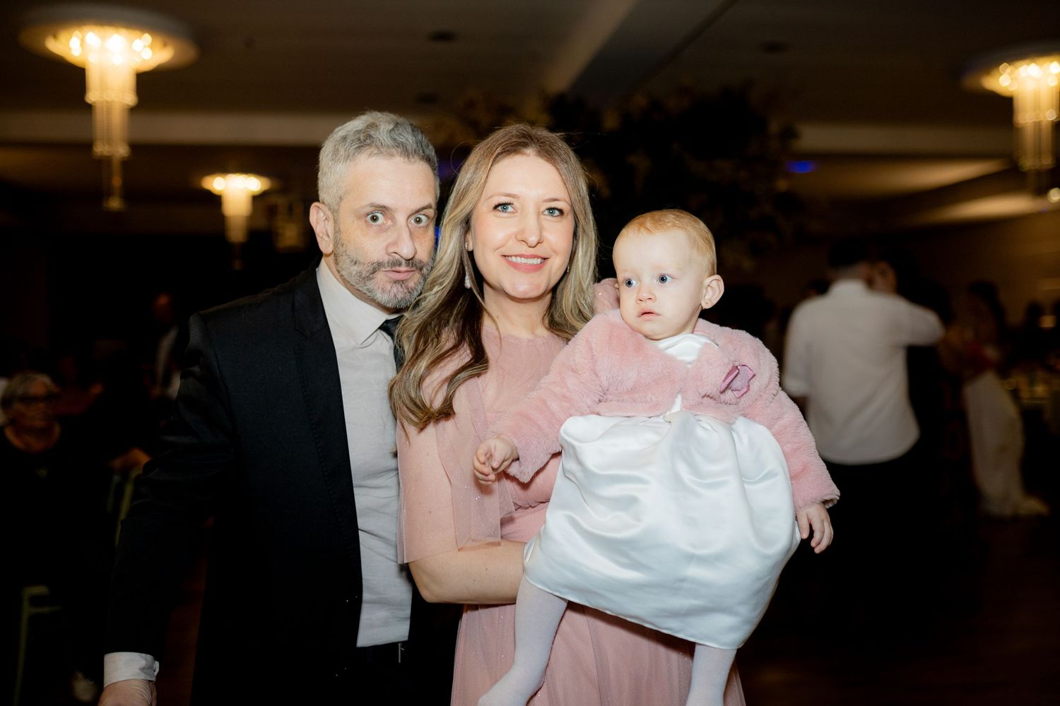 A couple in formal attire smiles while holding a baby dressed in white at an elegant event.