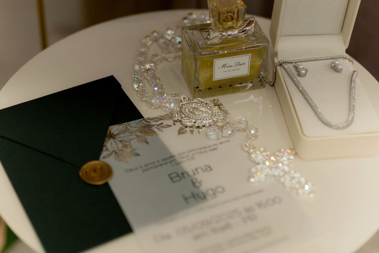 Elegant wedding invitation with gold wax seal, black envelope, perfume bottle, and pearl jewelry on white table.