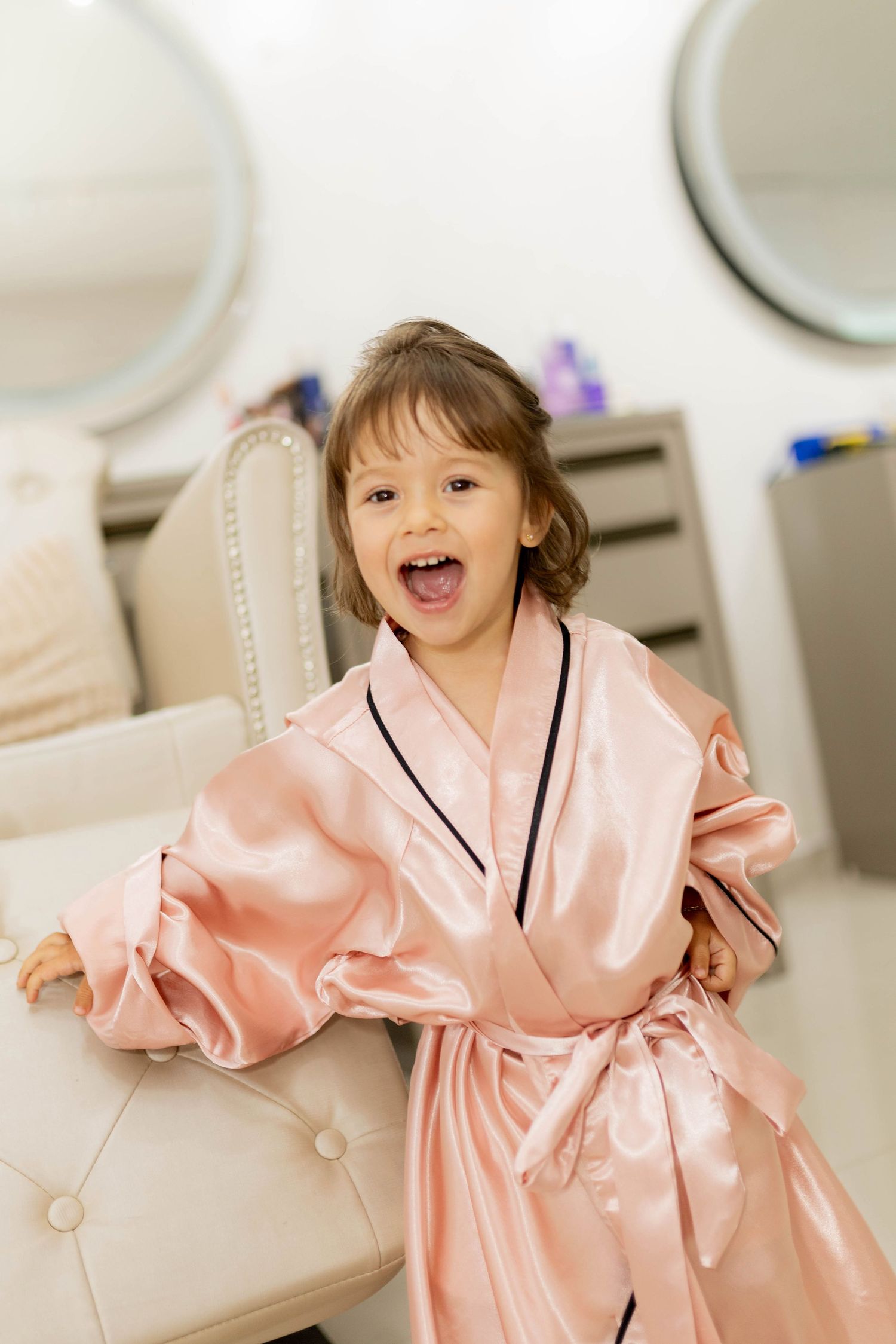 A joyful little girl in an oversized pink satin robe laughs while sitting in a beauty salon chair.