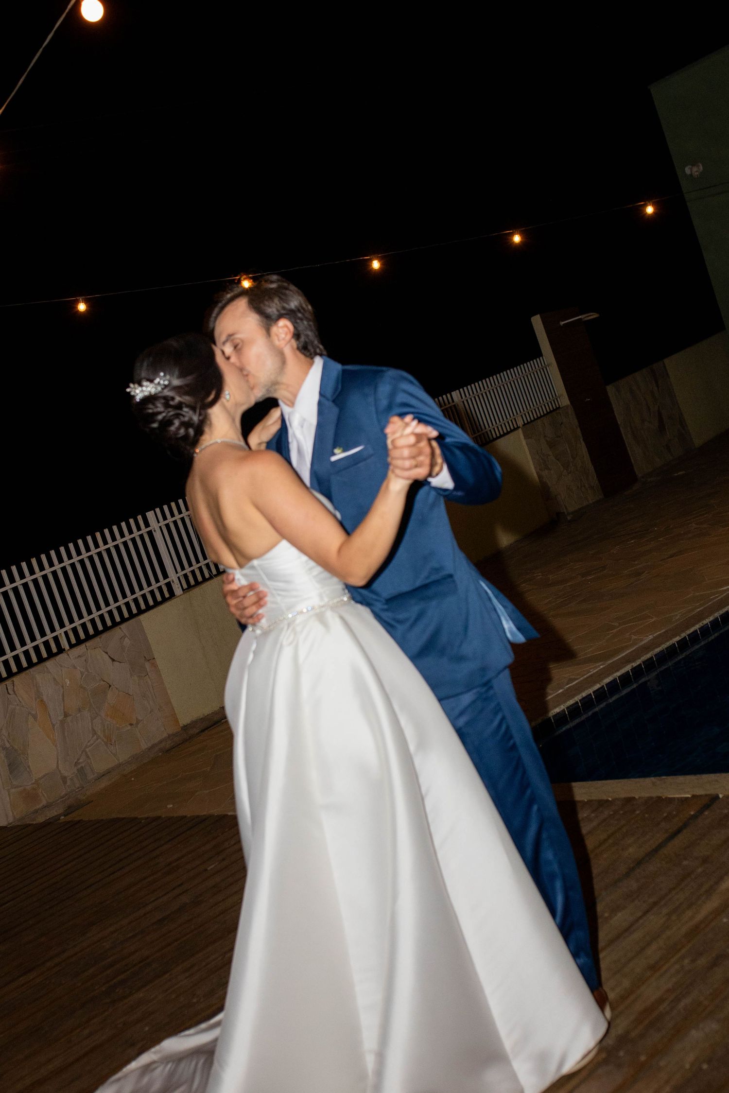 Bride and groom share first dance kiss outdoors at night, bride in white gown, groom in blue suit, string lights above.