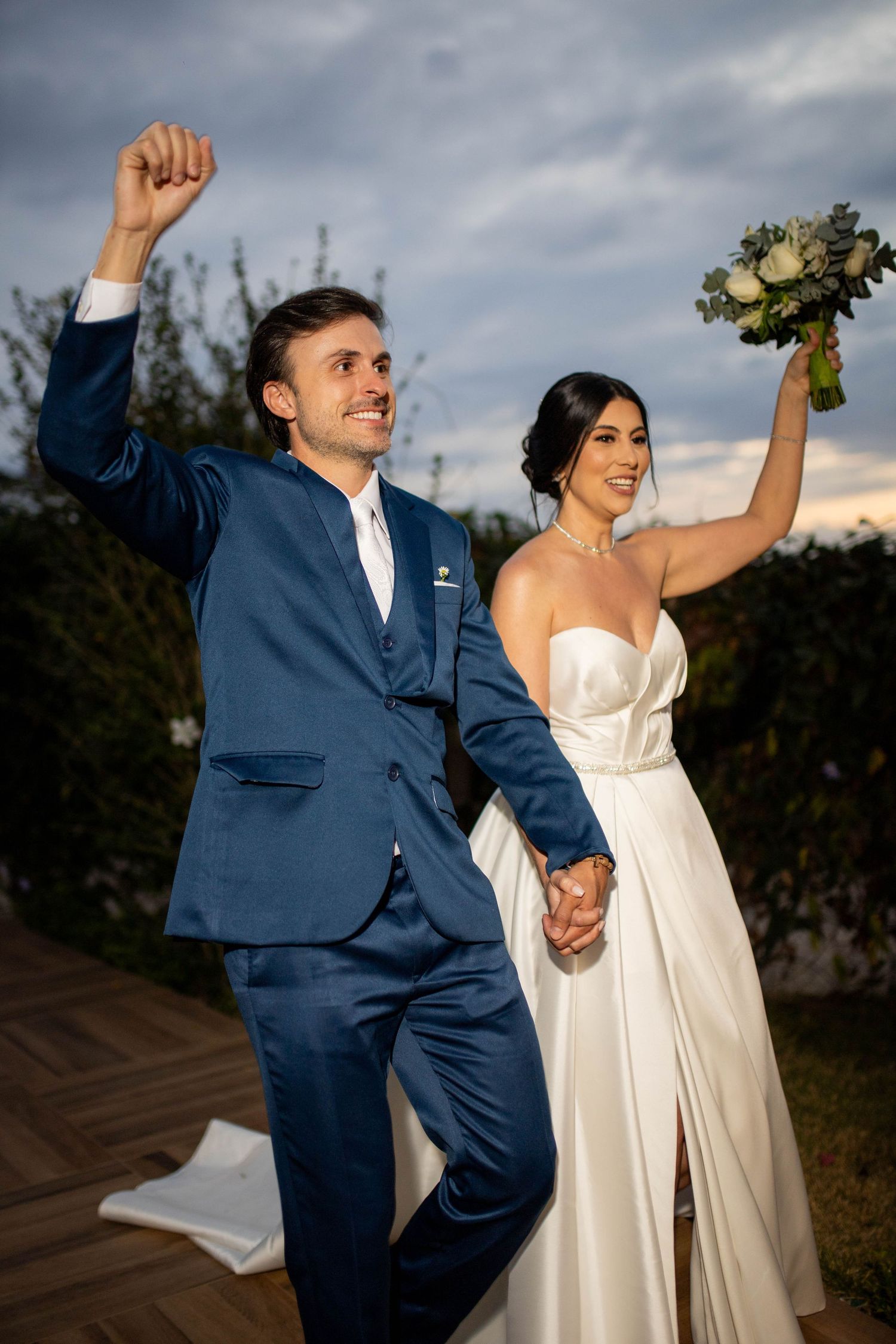 Joyful newlyweds celebrate outdoors, groom in blue suit raises fist, bride in white gown holds bouquet.