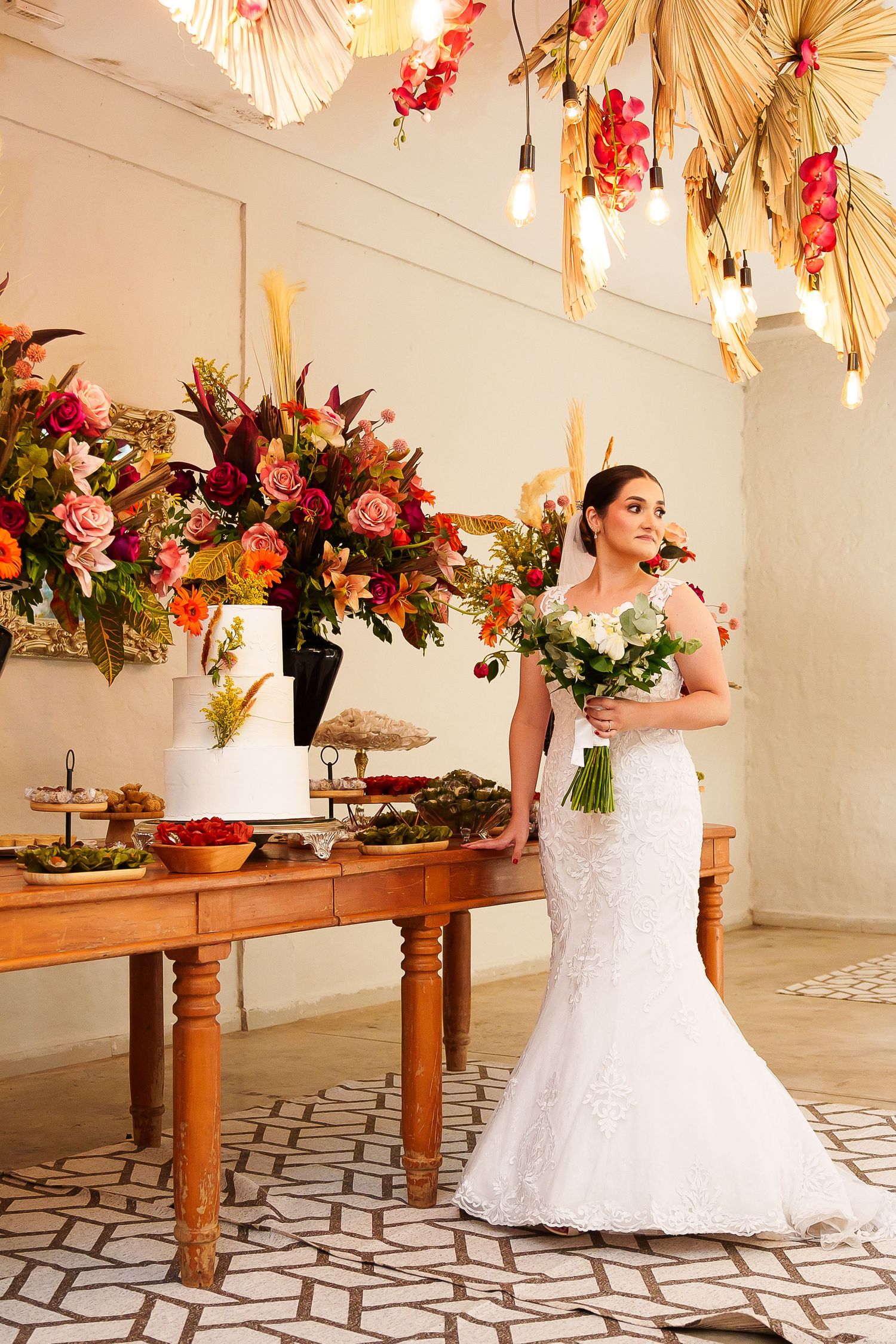 Bride in white lace mermaid gown holds bouquet beside floral wedding cake table with colorful arrangements and decorative lighting.