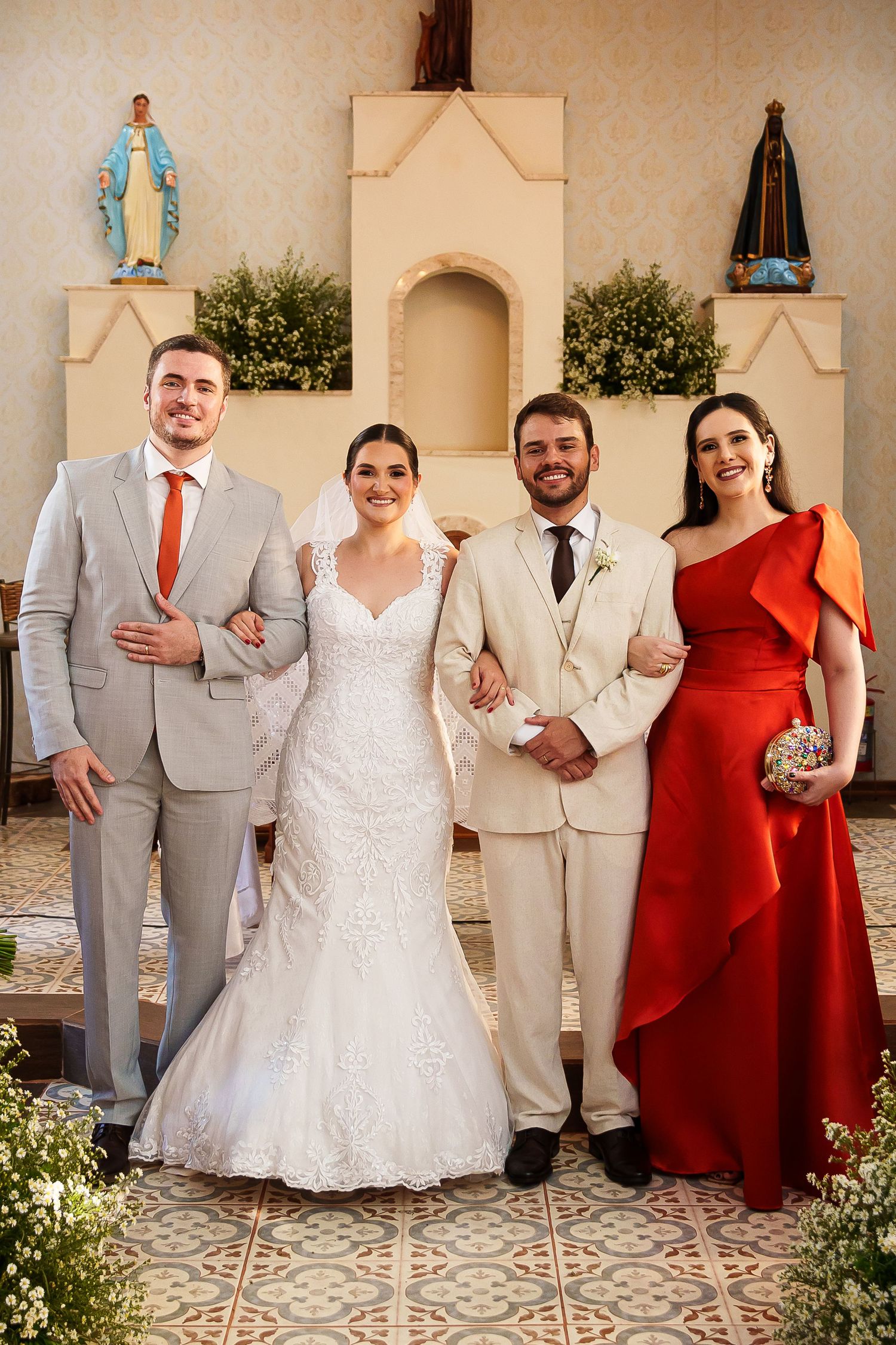 Wedding party poses in church, bride in white gown, groom in cream suit, groomsman in gray, bridesmaid in red dress.