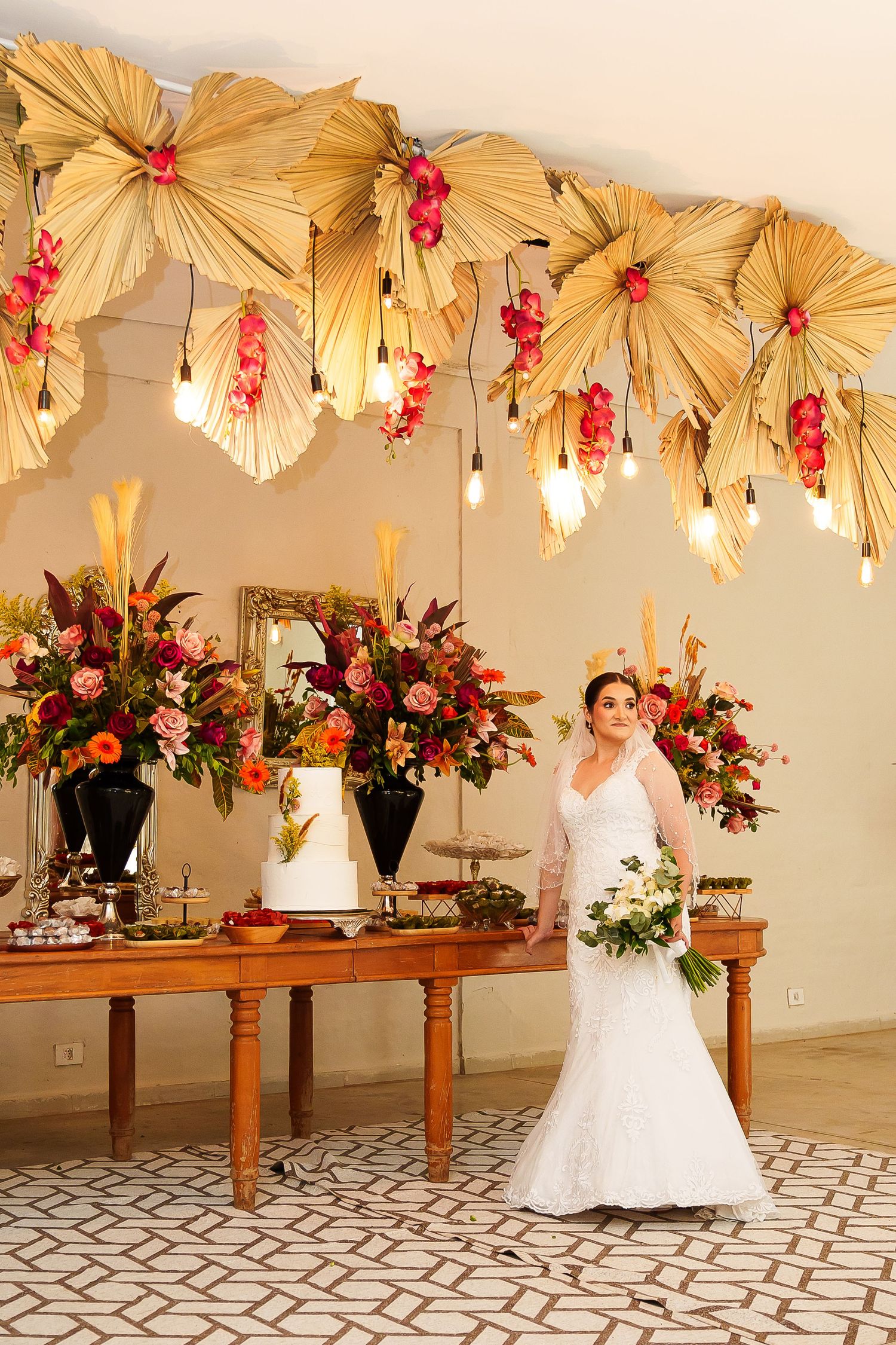 Bride in white lace gown holds bouquet beside floral wedding cake table with tropical palm leaf ceiling decor.