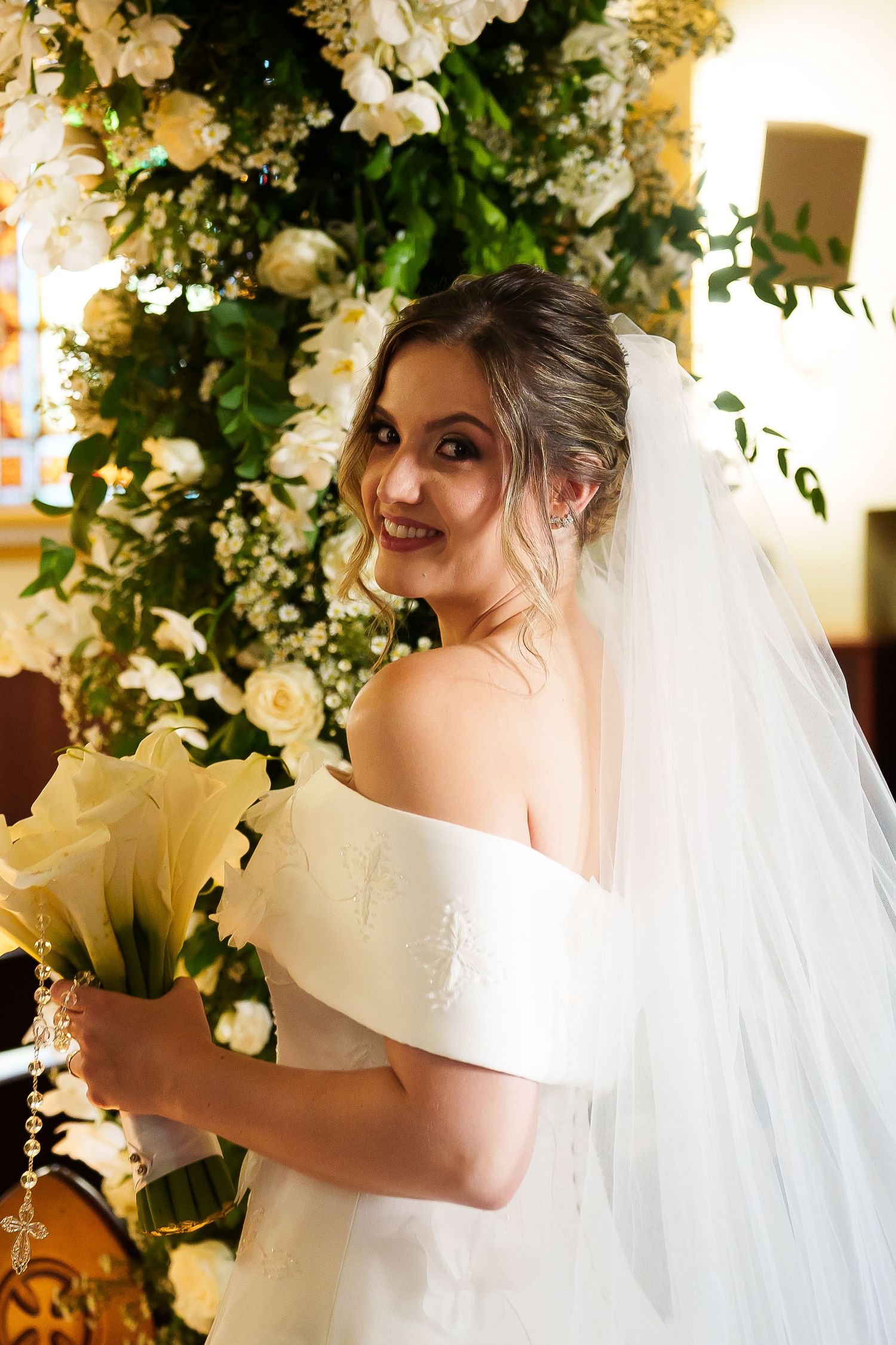Smiling bride in off-shoulder white gown and veil holds yellow lily bouquet before lush floral arrangement.