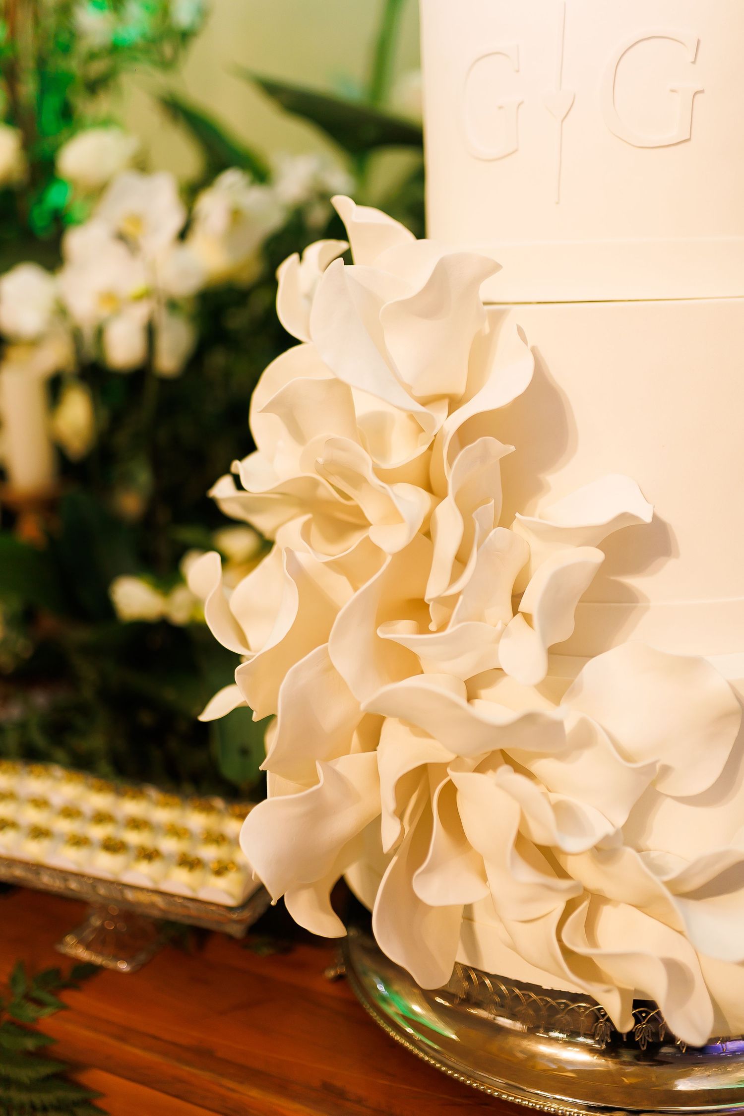 Elegant white wedding cake with large ruffled sugar flowers and monogram initials, displayed on a silver stand.