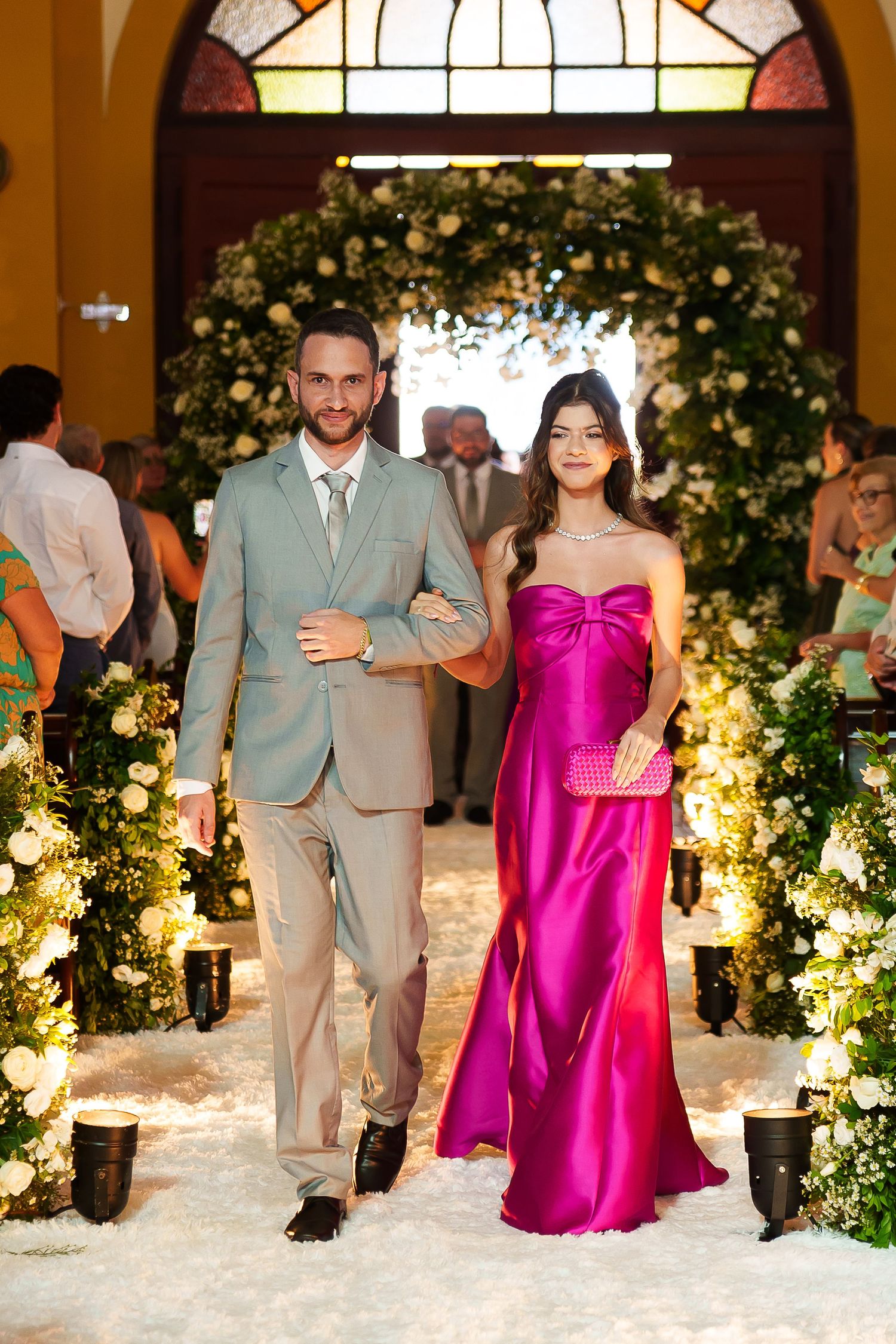 Couple walking down wedding aisle, man in gray suit, woman in strapless magenta gown, floral arch backdrop.