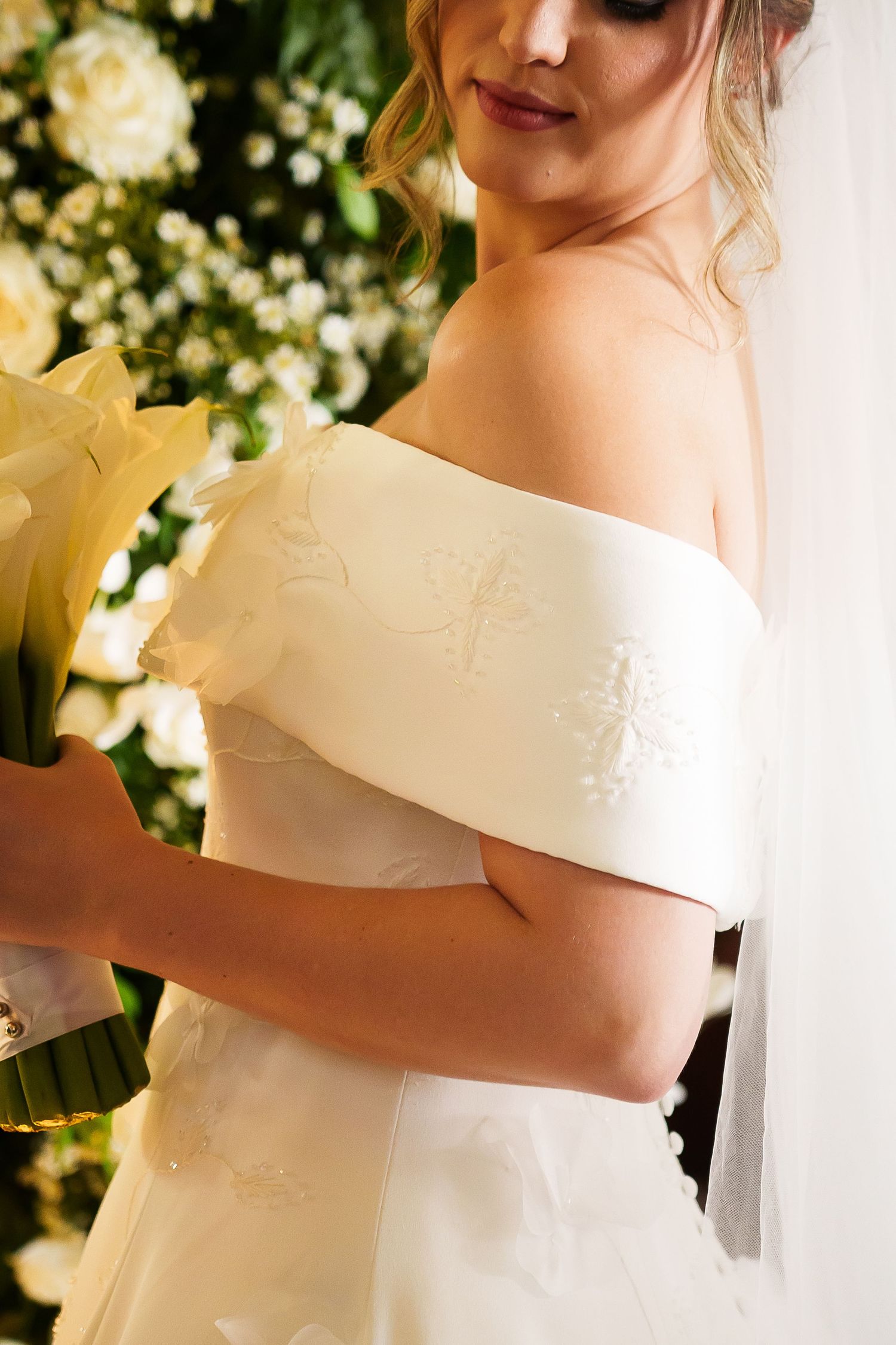 Bride in off-shoulder white gown with floral embroidery holds yellow lily bouquet near white flower arrangement.