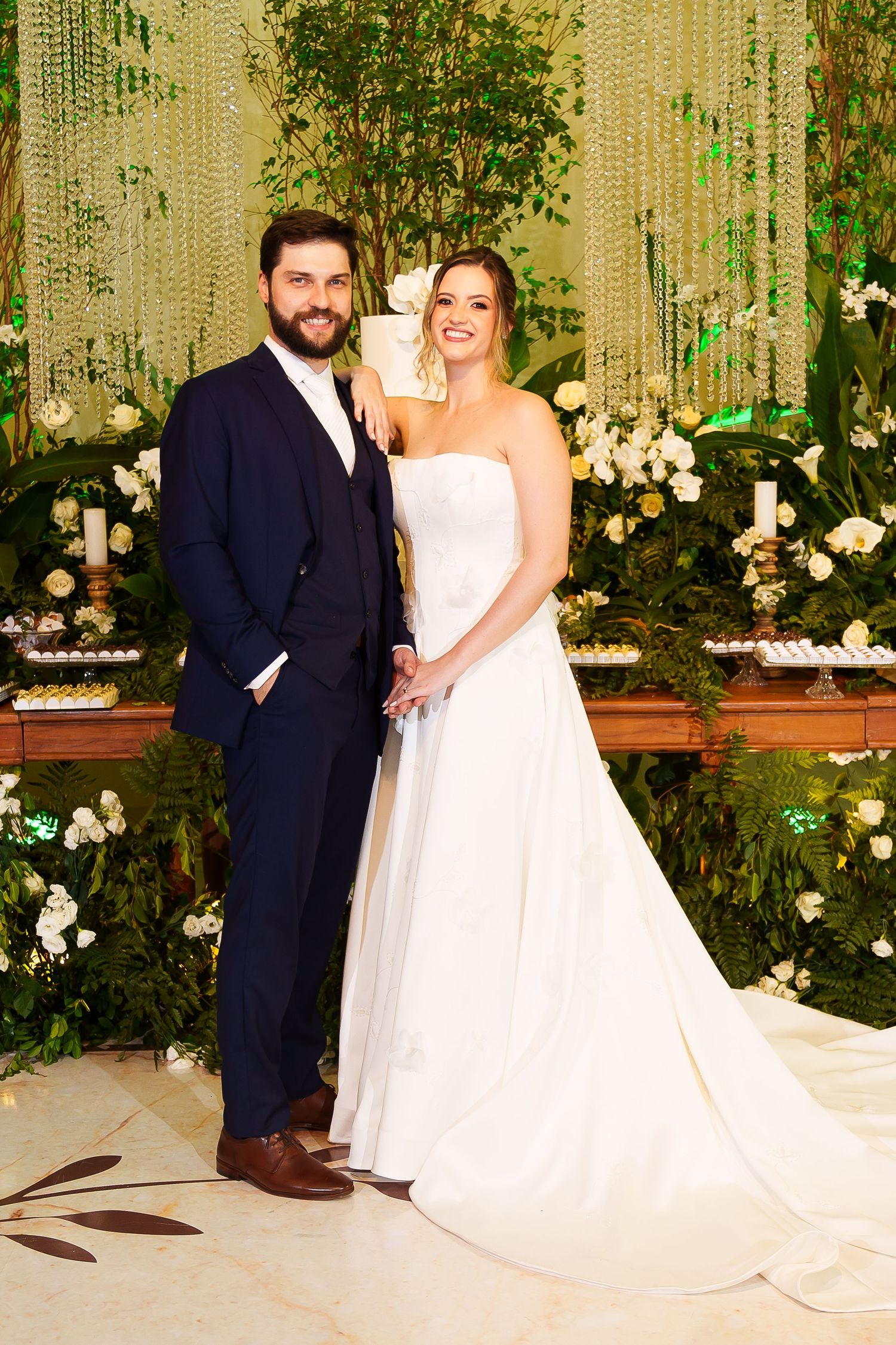Bride and groom posing together at wedding reception with lush green floral backdrop and white roses.