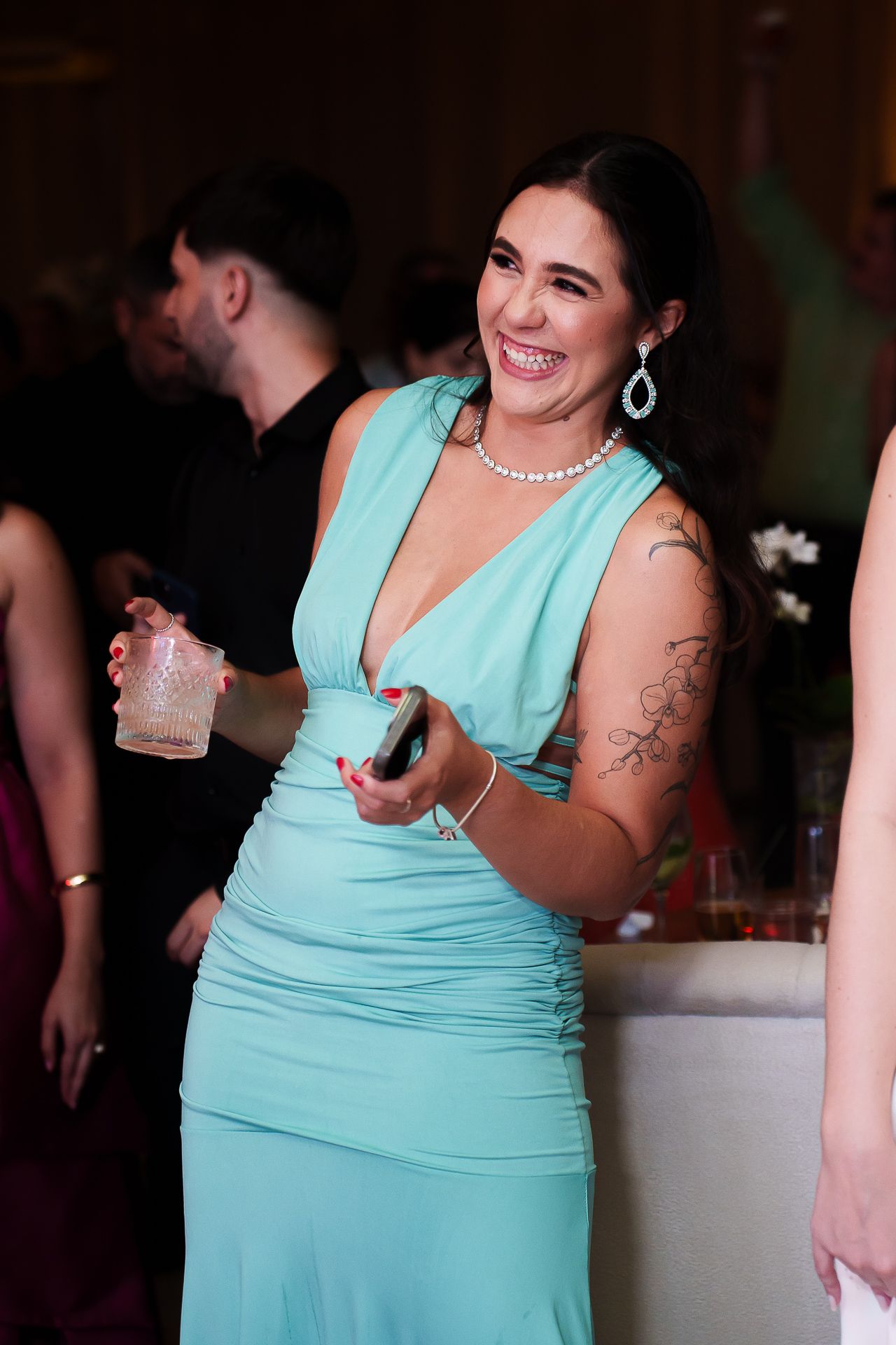 Woman in teal gown with tattoos smiling at formal event, wearing pearl necklace and holding drink.