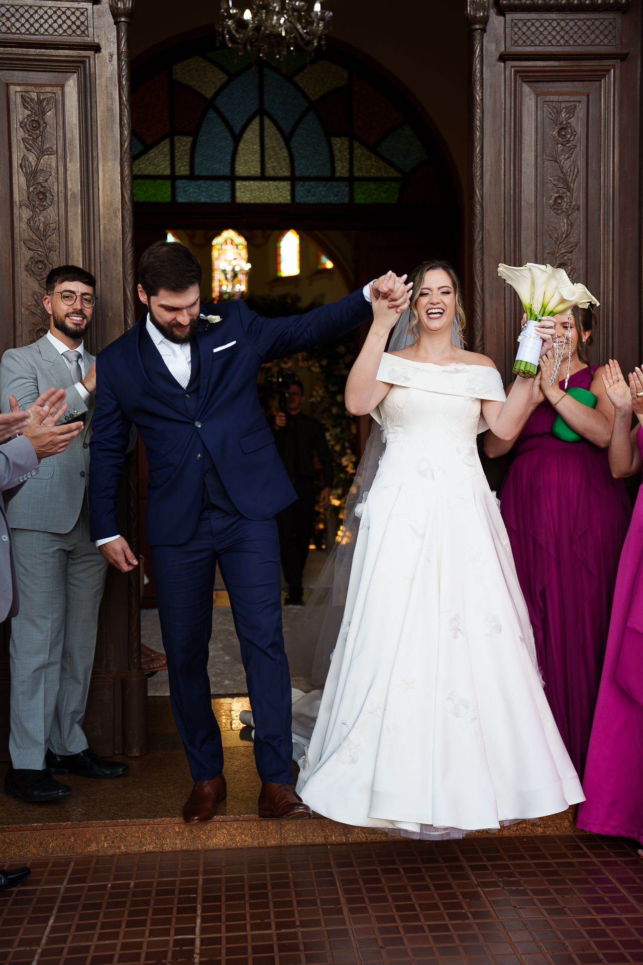 Joyful newlyweds exit church doors, bride in white gown holding bouquet, groom in navy suit, guests applauding around them.