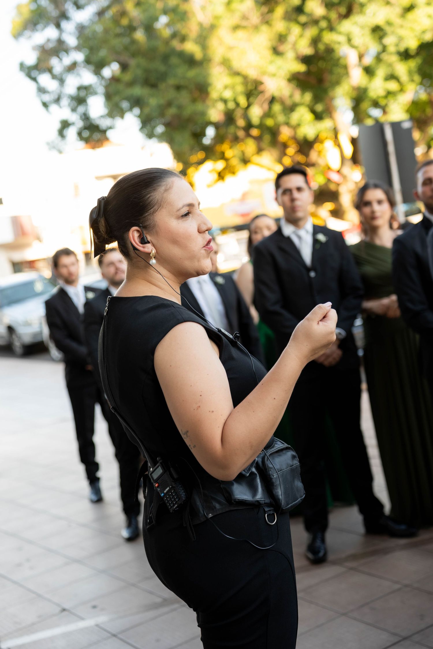 Woman in black outfit with earpiece and bag stands before group in formal attire outdoors.