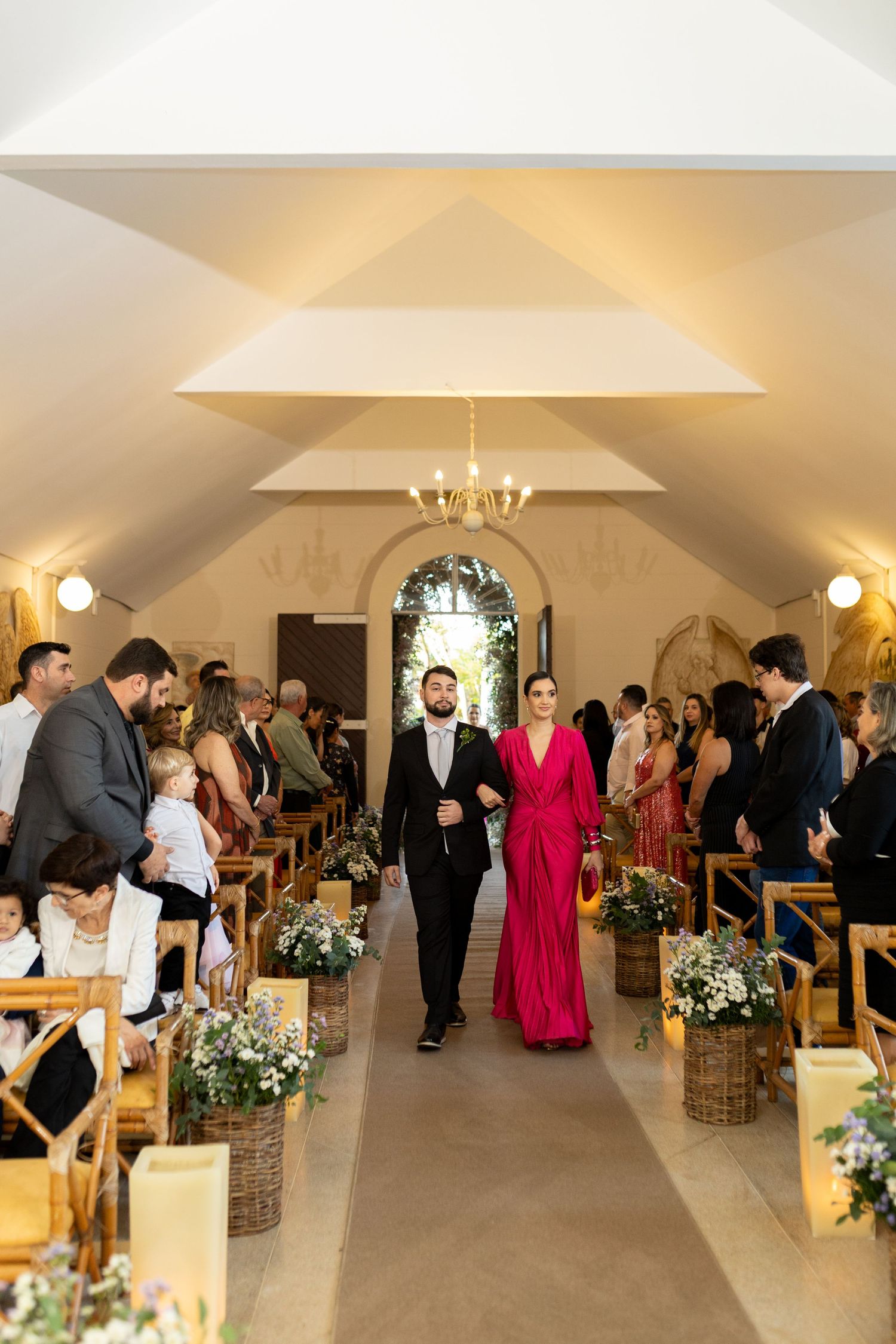 A groom in a black tuxedo escorts a woman in a red gown down a floral-decorated church aisle at a wedding.