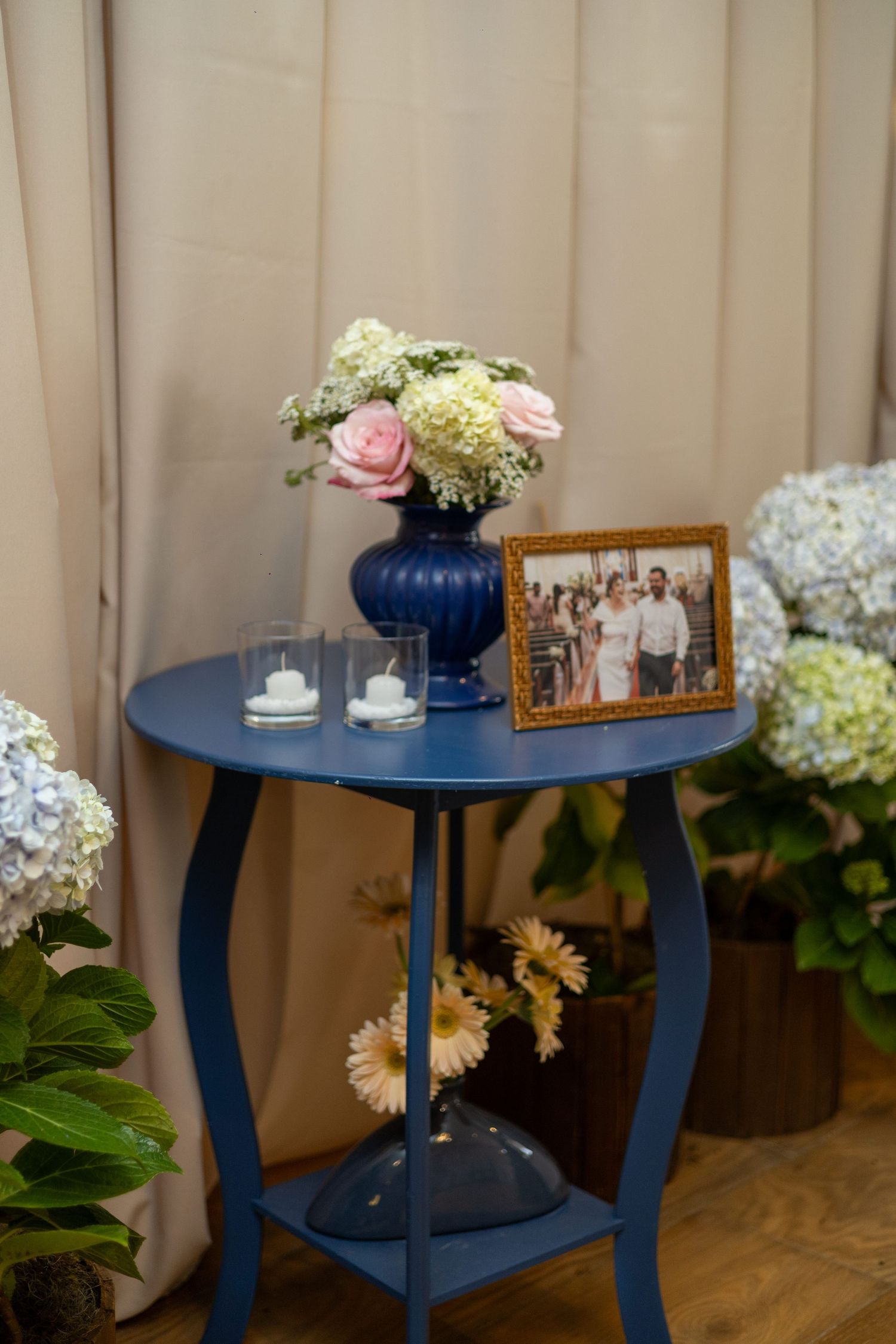 Blue side table with floral vase, candles, and framed photo, surrounded by hydrangeas in elegant decor.