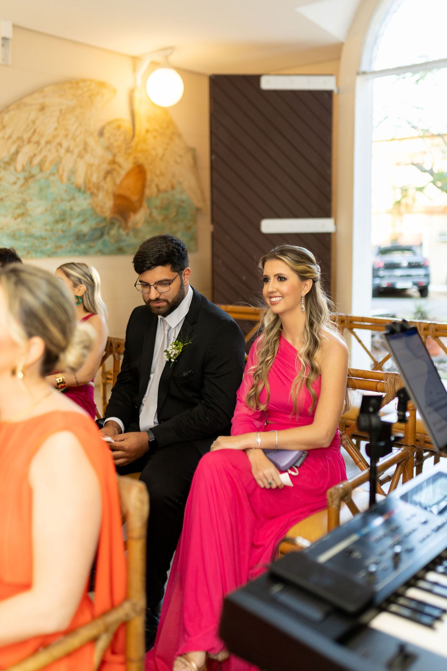 Guests in formal attire seated at an indoor wedding ceremony, woman in pink dress smiling near a keyboard.