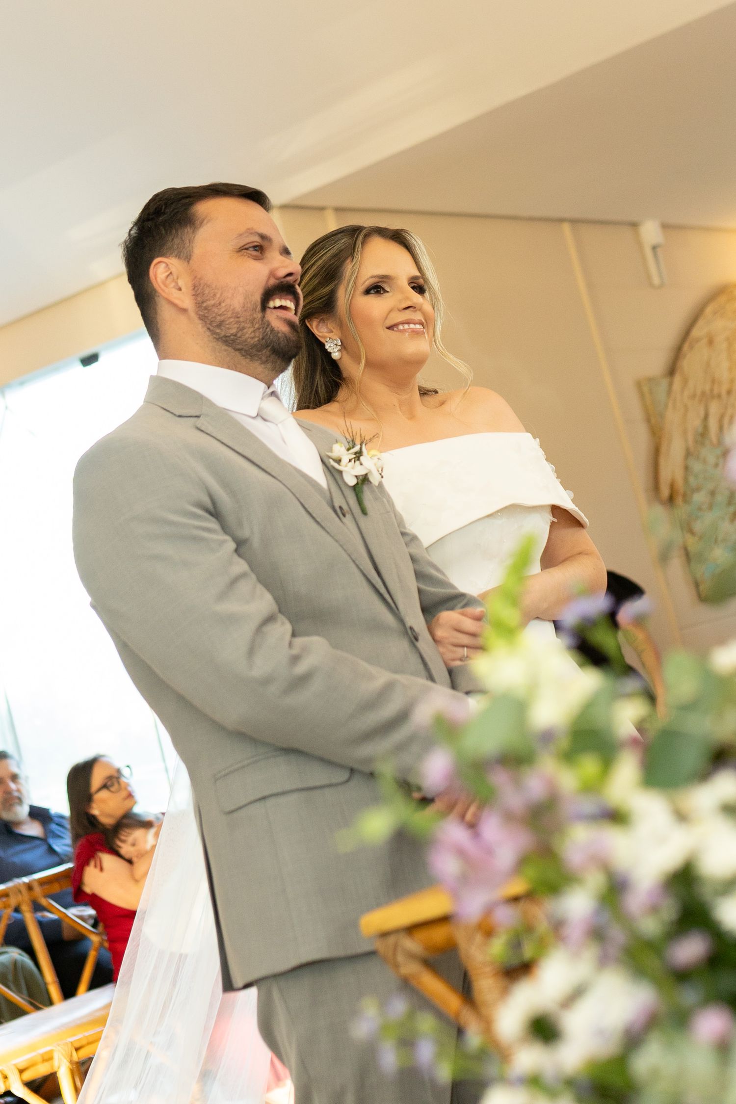 A smiling bride in a white off-shoulder gown and groom in a gray suit stand together at their wedding ceremony altar.