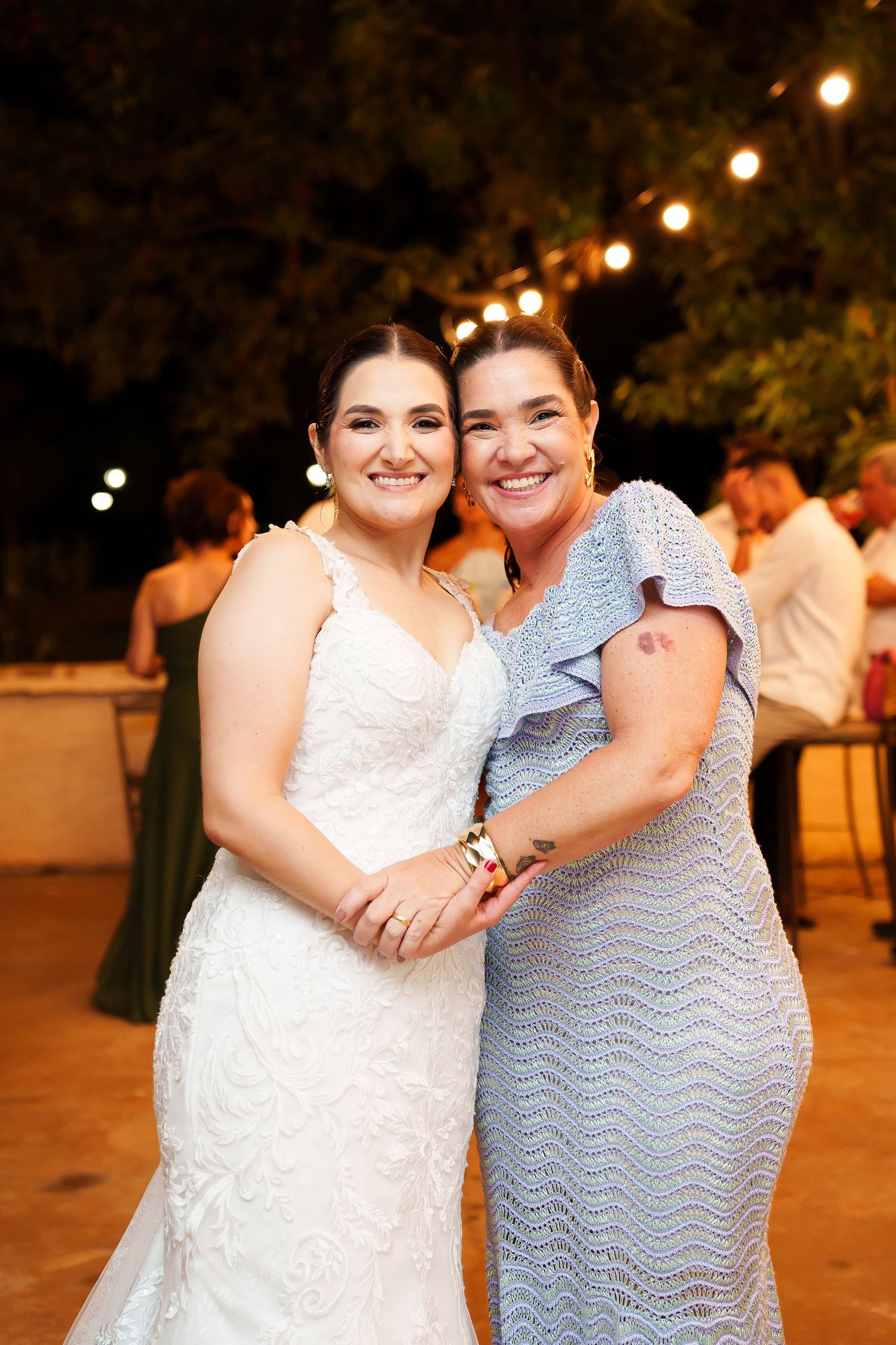 Bride in white lace gown poses with smiling woman in blue dress at outdoor evening wedding reception.