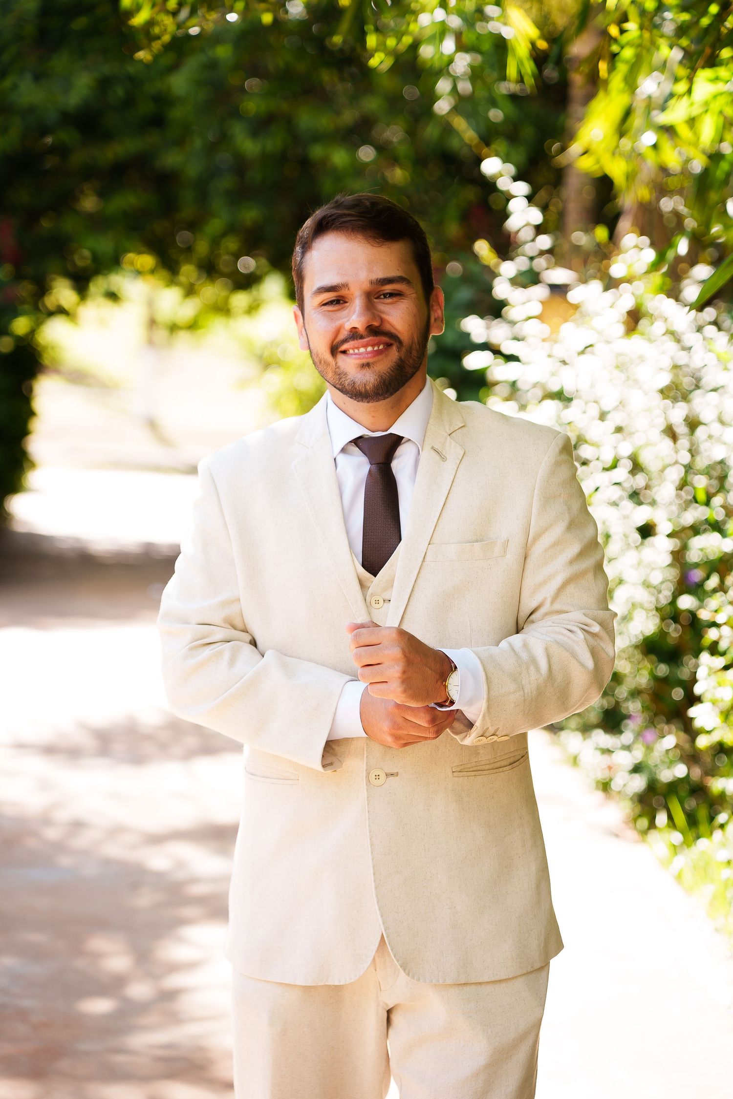 Smiling groom in cream suit and brown tie adjusting cufflinks outdoors among lush greenery.