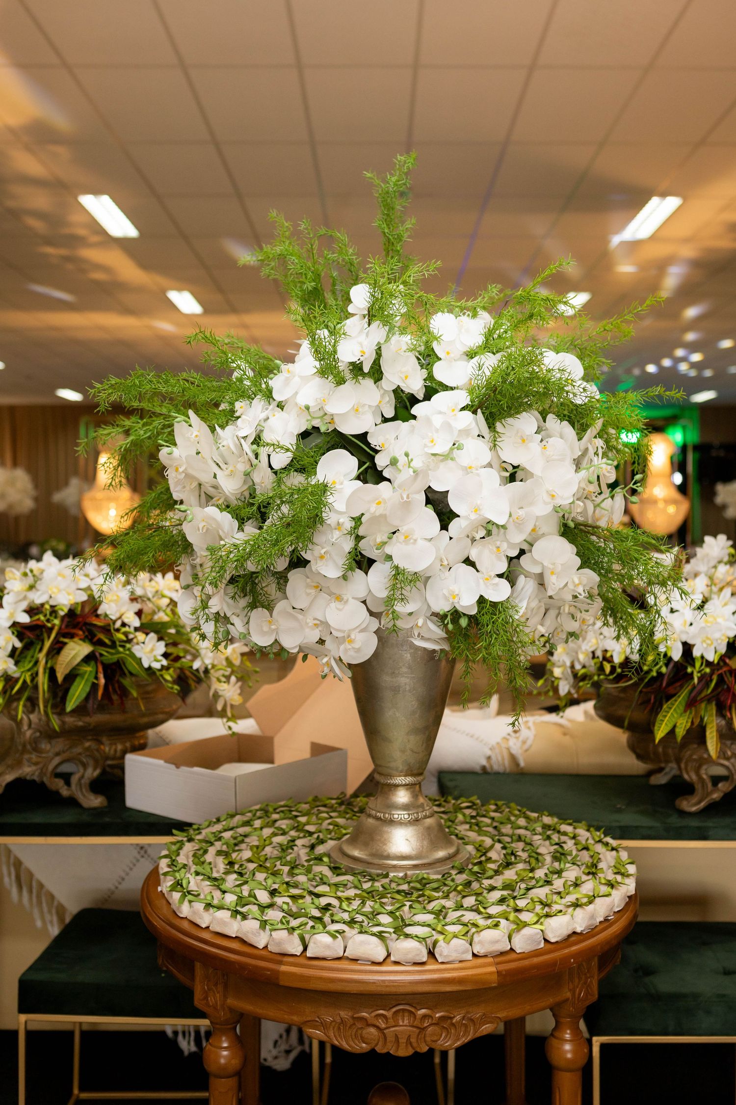 Elegant silver vase with white orchids and green ferns on a decorative round table in a wedding venue.