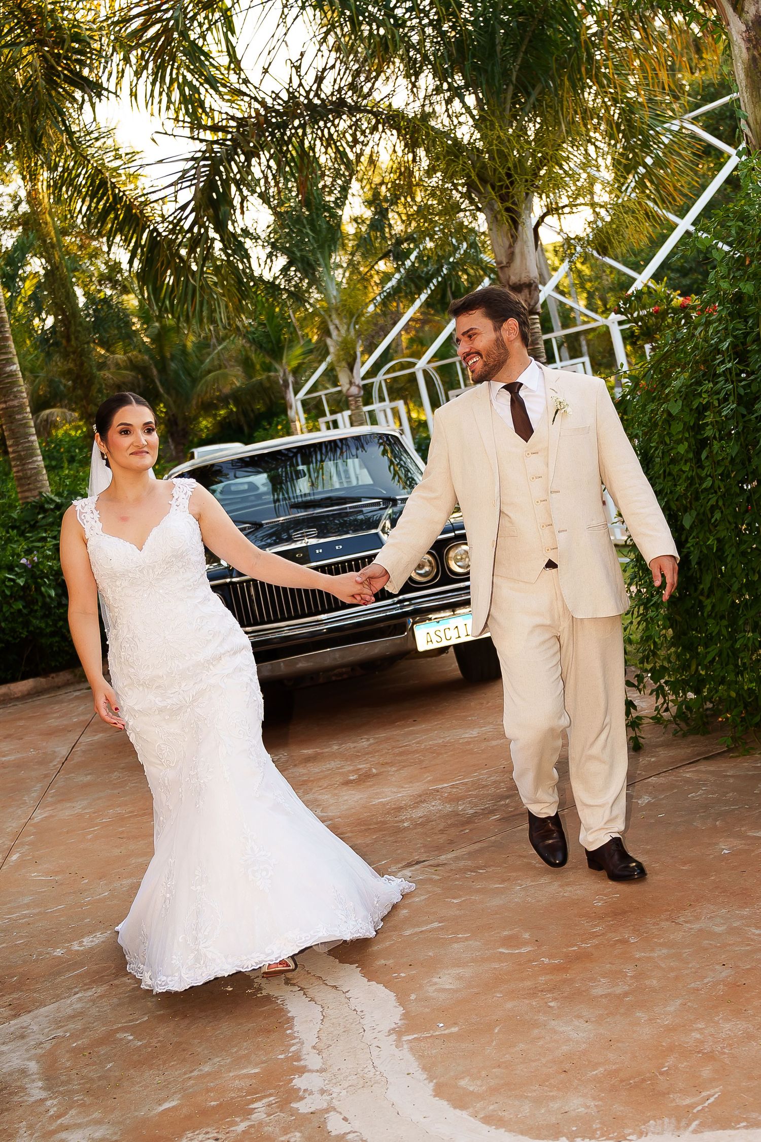 Bride in white lace gown and groom in beige suit hold hands walking past a classic black car amid tropical greenery.