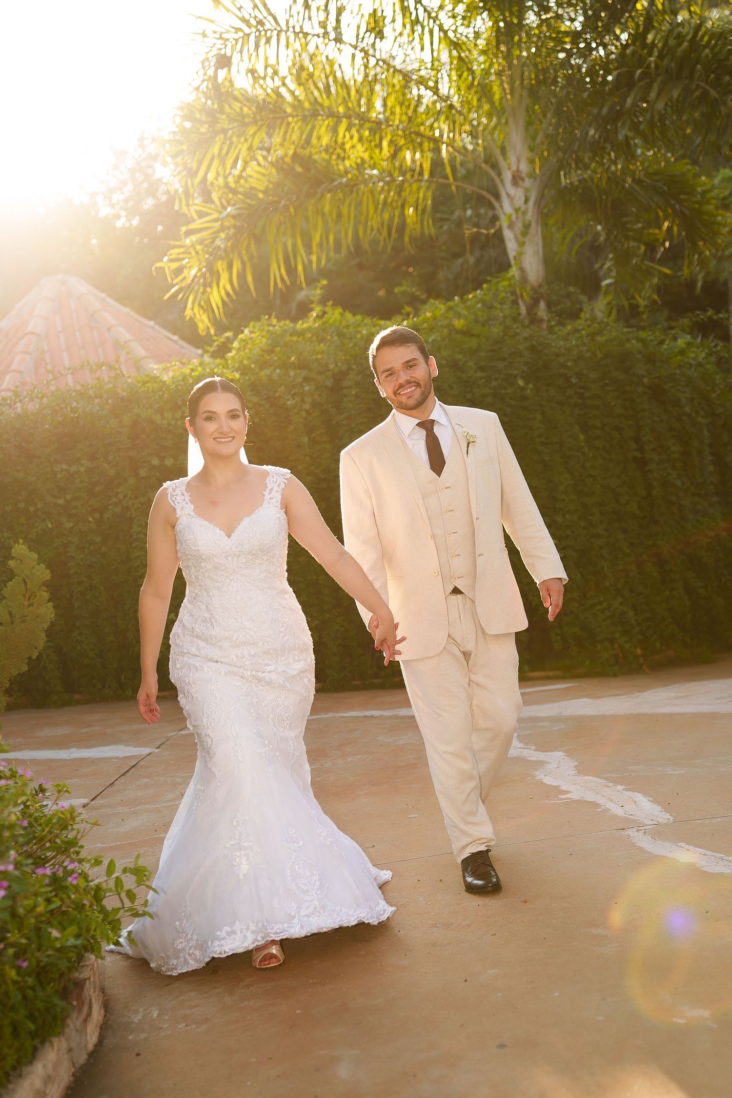 Bride and groom walk hand in hand outdoors in golden sunset light, she in white lace gown, he in cream suit.