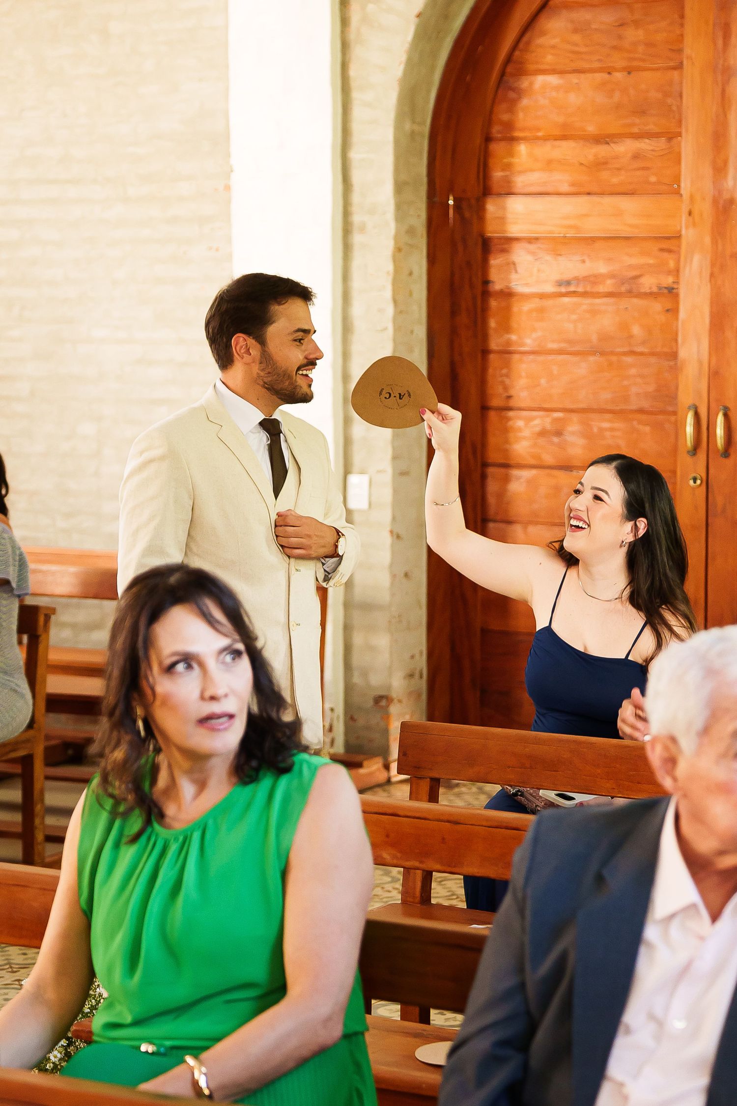 Guests laughing and socializing inside a church before a wedding ceremony, with wooden pews and arched doorway.