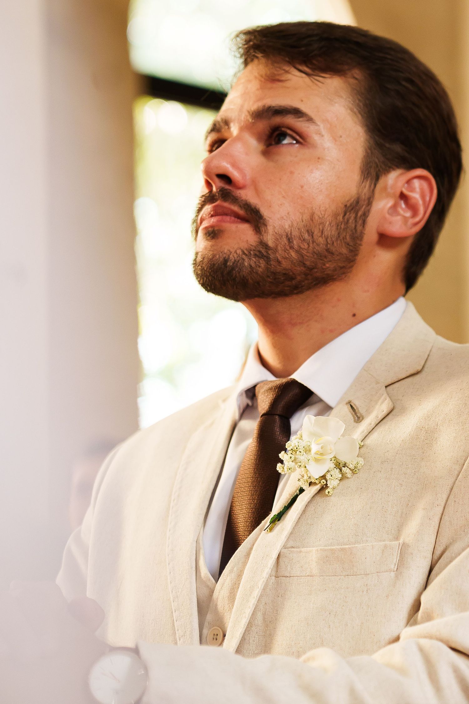 Bearded groom in beige suit with white boutonniere gazing upward at wedding ceremony.