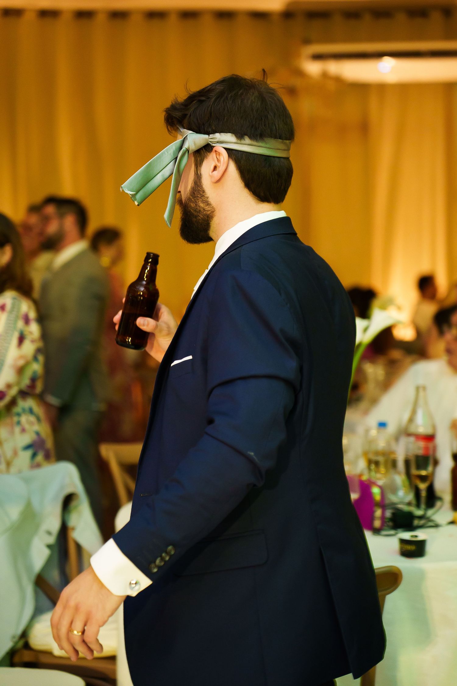 Man in navy suit with ribbon blindfold holding beer bottle at elegant wedding reception party.