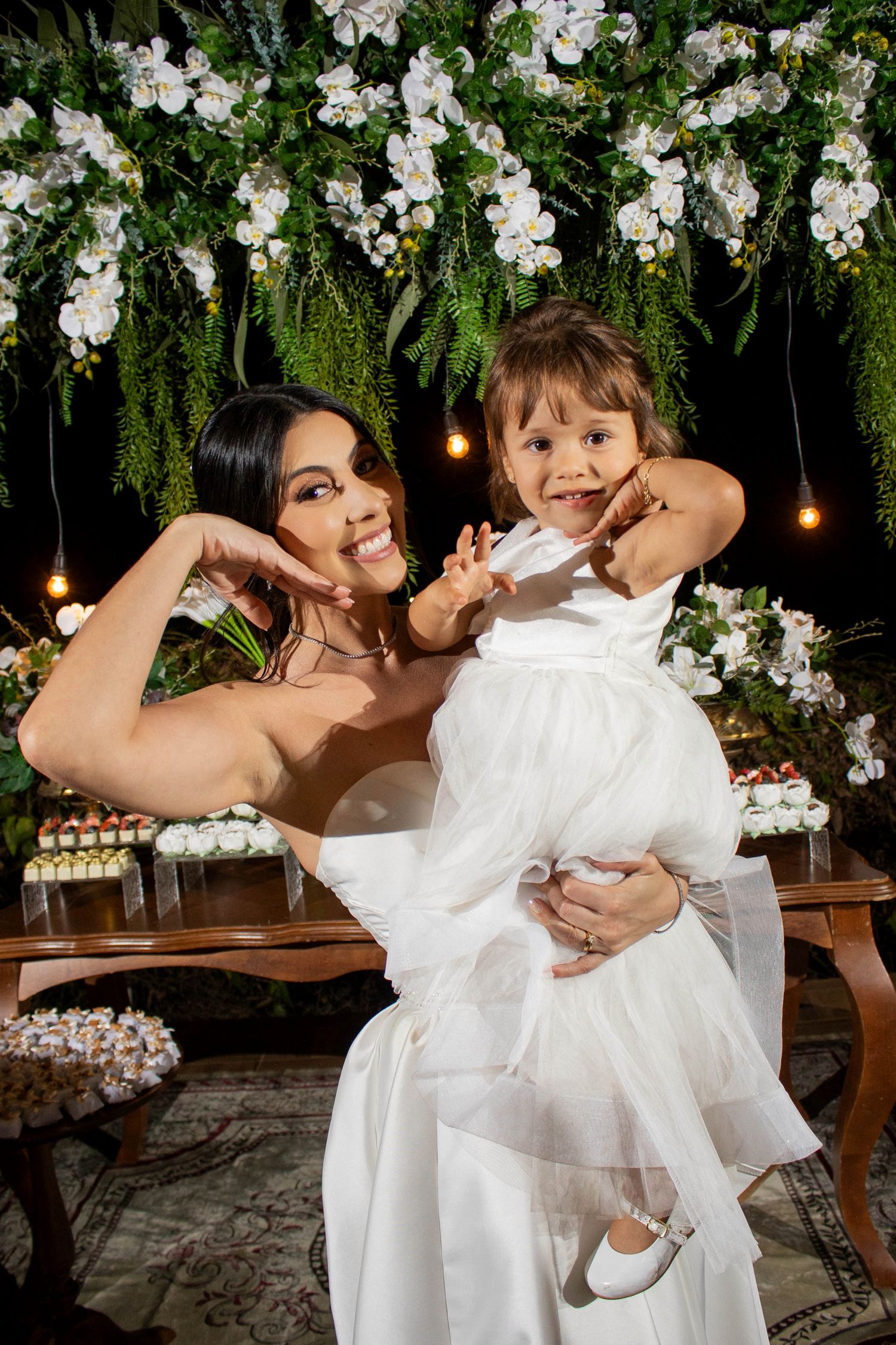 Woman in white dress holds smiling toddler girl in white dress before floral backdrop at elegant celebration.