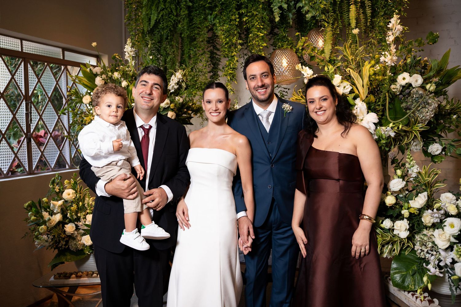 Wedding party poses with bride and groom in front of floral greenery wall, including family members and a toddler.