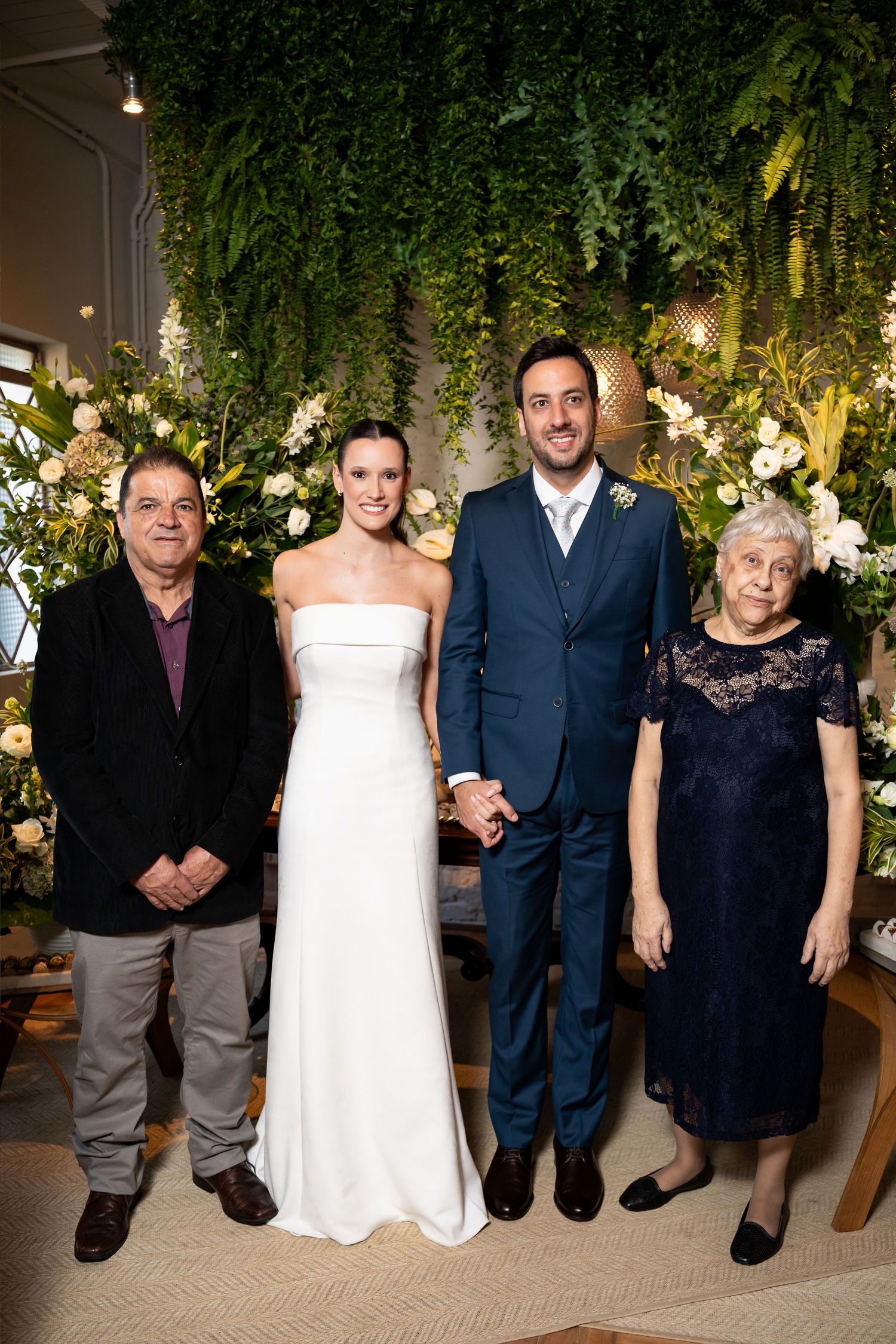 Bride and groom pose with two elderly guests in front of lush green floral backdrop at elegant wedding ceremony.