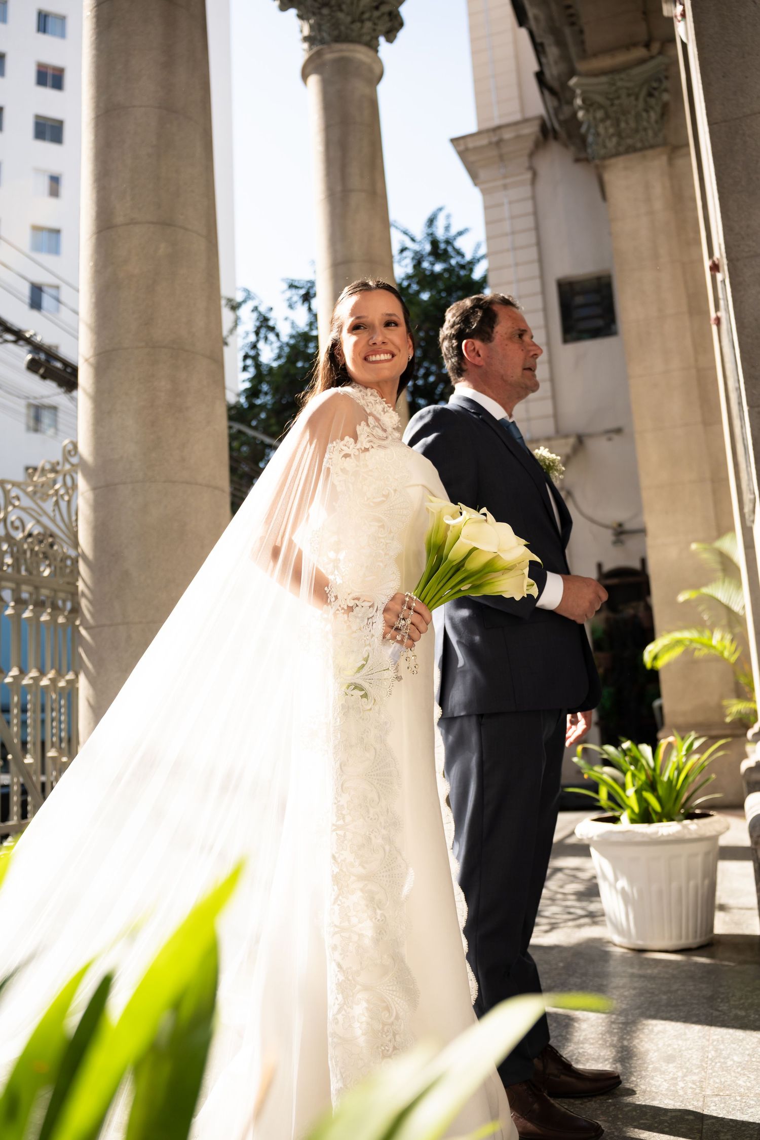 Bride in white lace gown holding calla lilies stands with groom in navy suit on church steps.