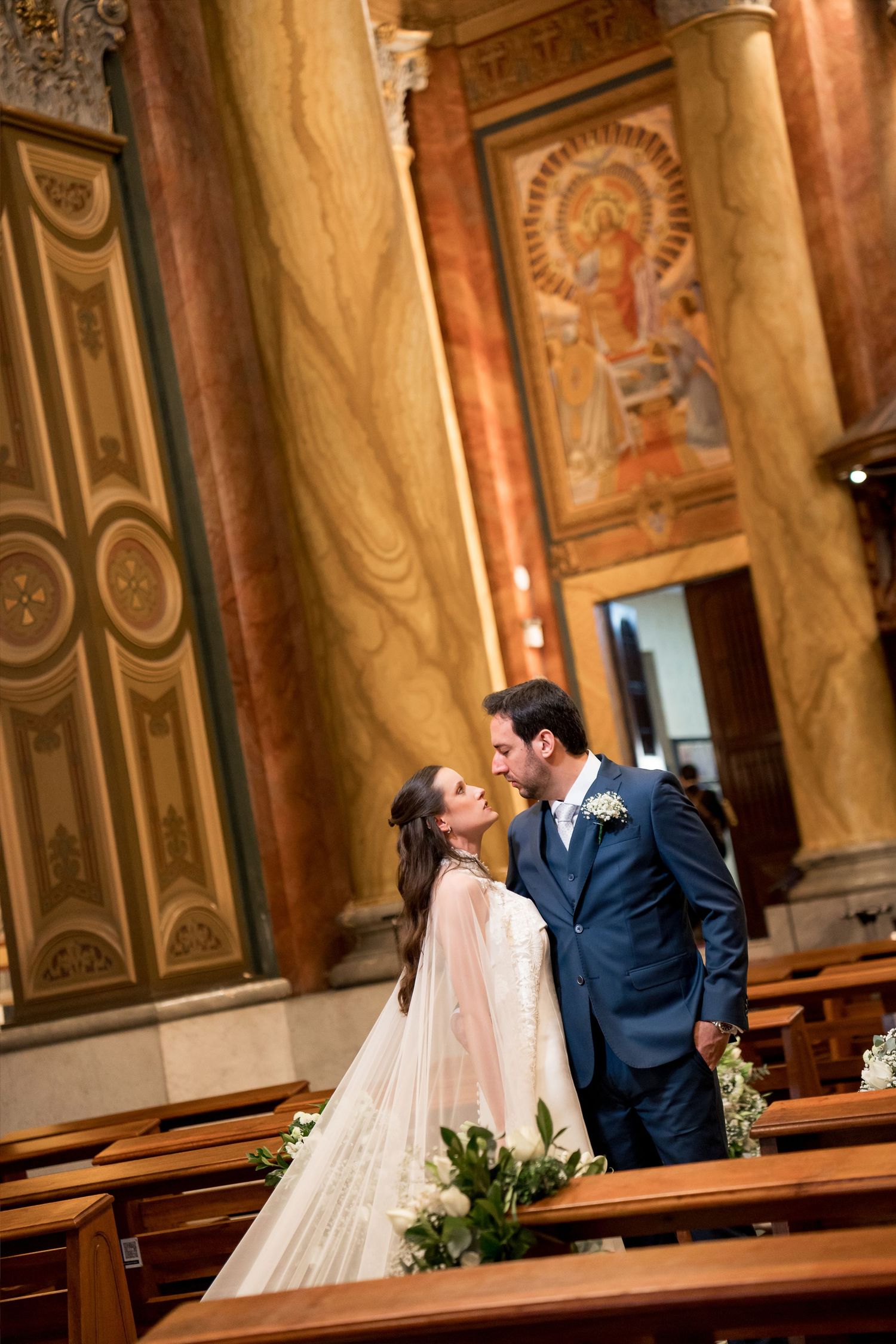 Bride and groom share a romantic kiss in an ornate church with marble columns and mosaic artwork.