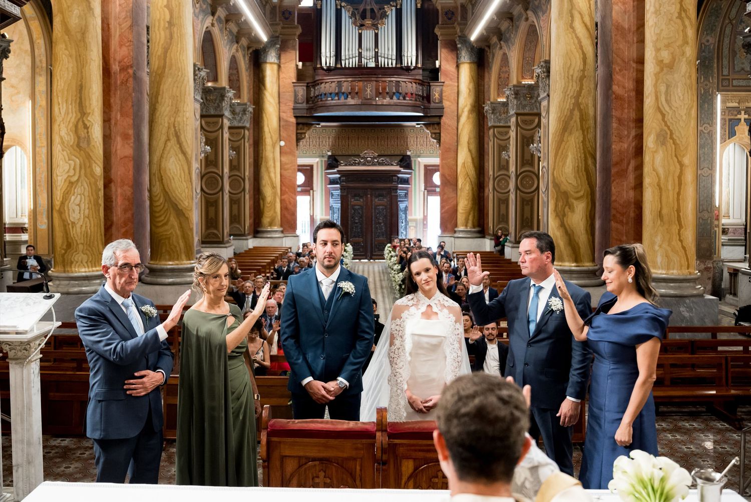 Elegant church wedding ceremony with couple at altar, flanked by family members in formal attire, ornate marble interior.