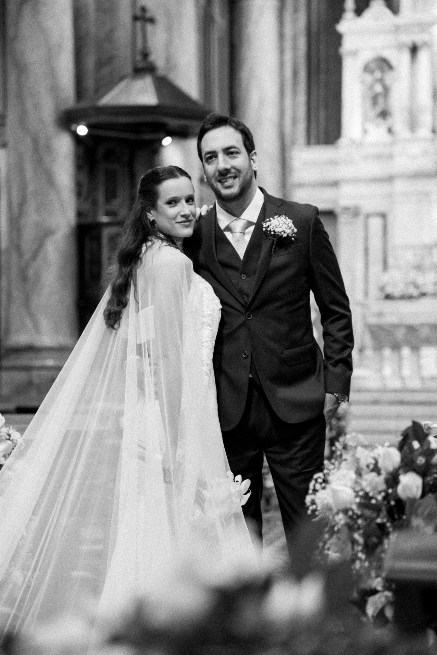 Black and white wedding photo of smiling bride in veil and groom in suit posing inside a church.