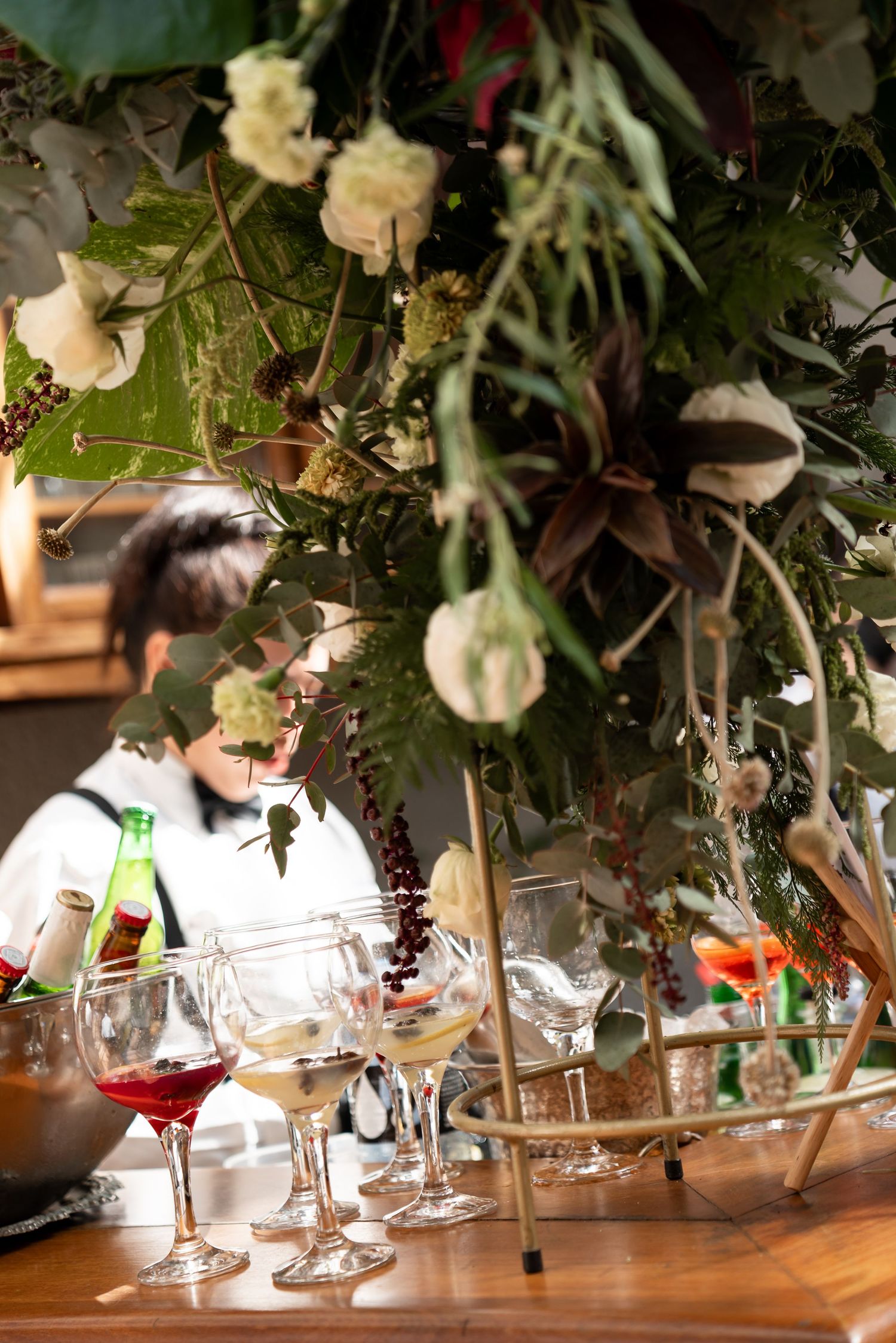Elegant wine glasses and floral arrangement on a wooden bar with a bartender in the background.