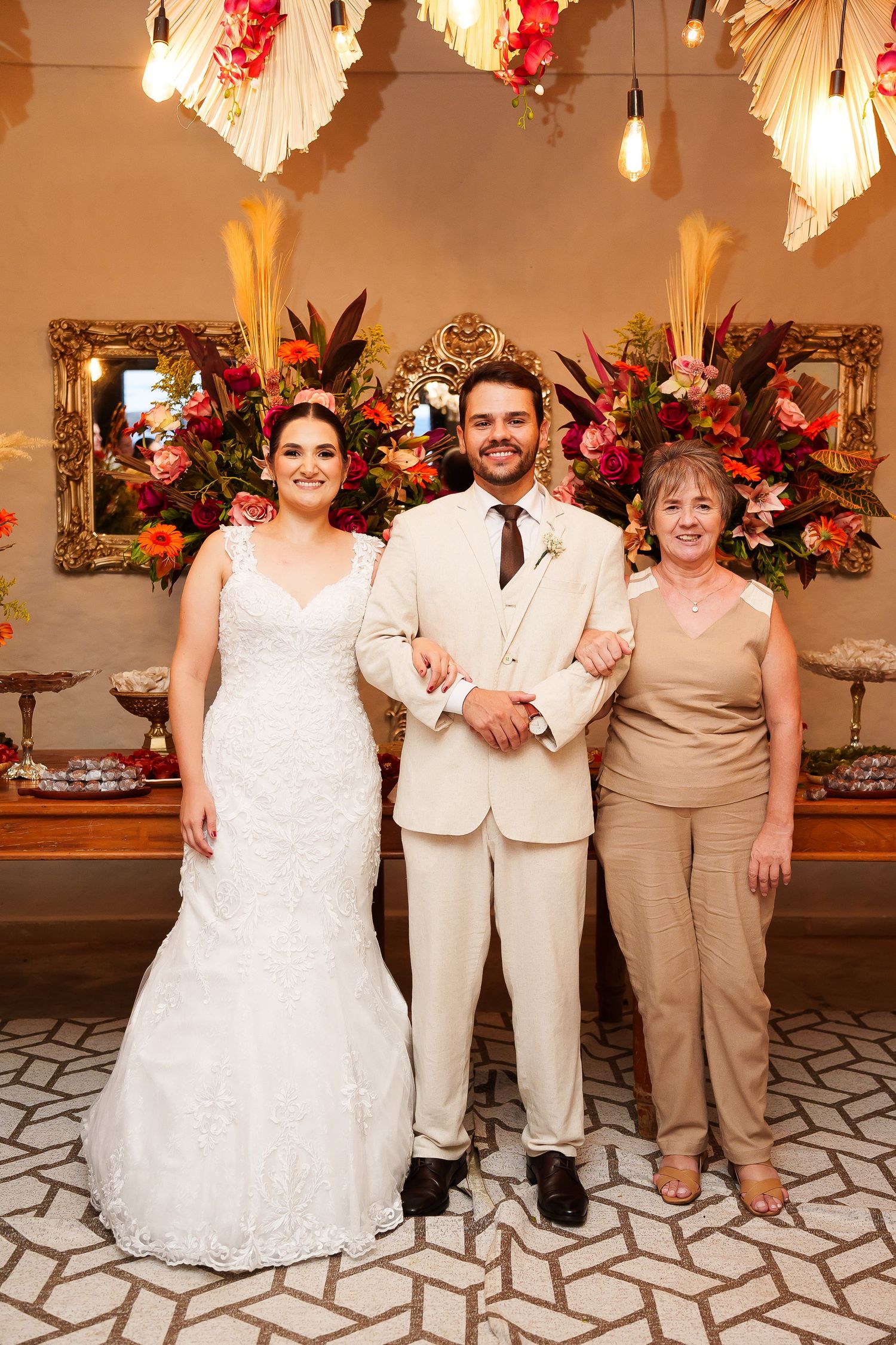 Bride in white gown and groom in beige suit pose with older woman at elegant floral wedding reception.