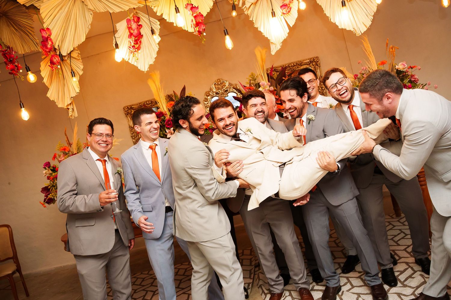 Groomsmen in gray suits lift the groom during a fun wedding party celebration indoors.