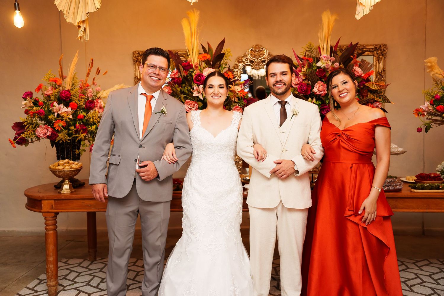 Wedding party photo with bride in white gown, groom in cream suit, and two guests in gray and red attire.