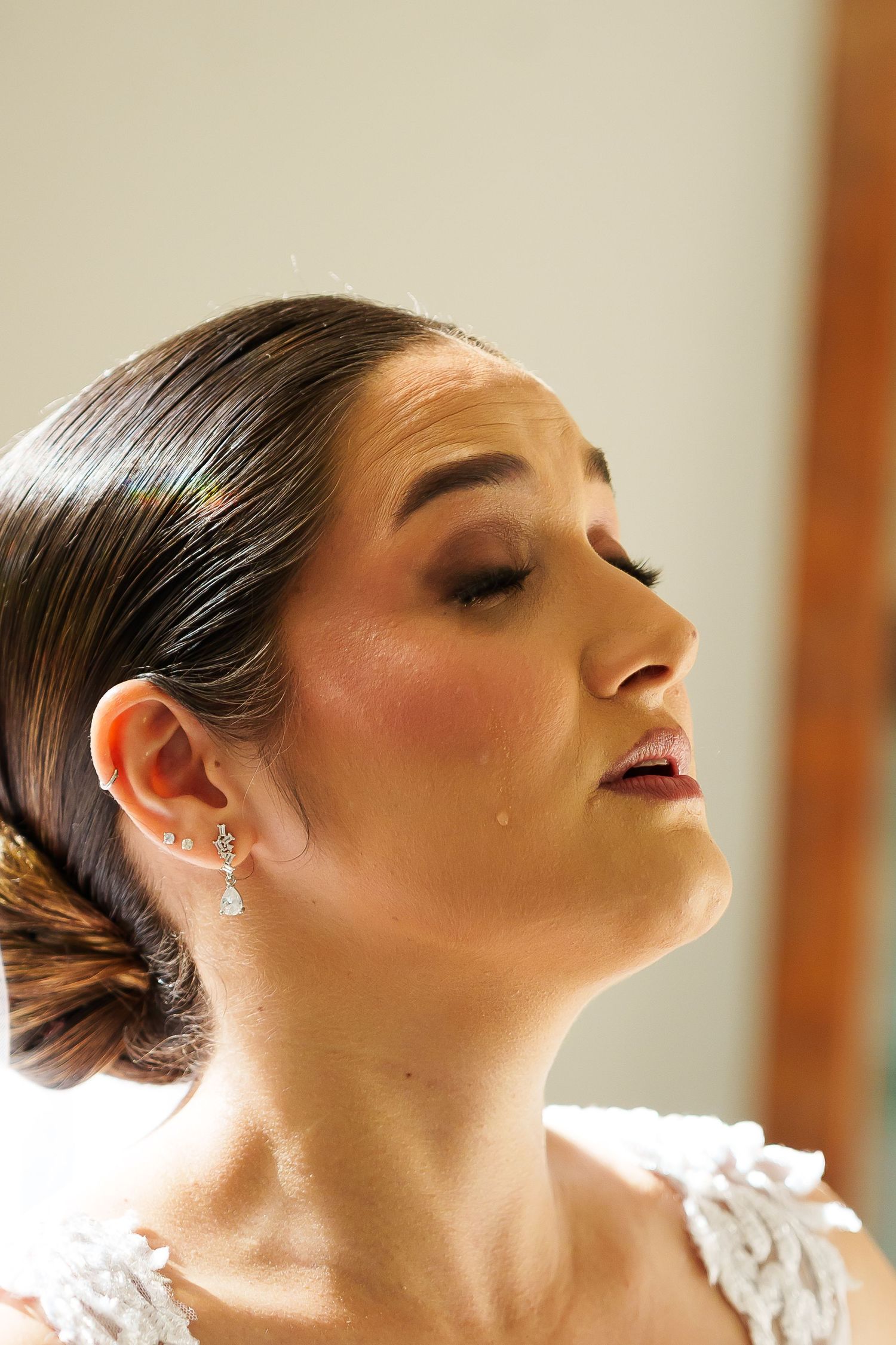 Bride with sleek updo and teardrop earrings, eyes closed, shedding a tear in her white lace wedding dress.
