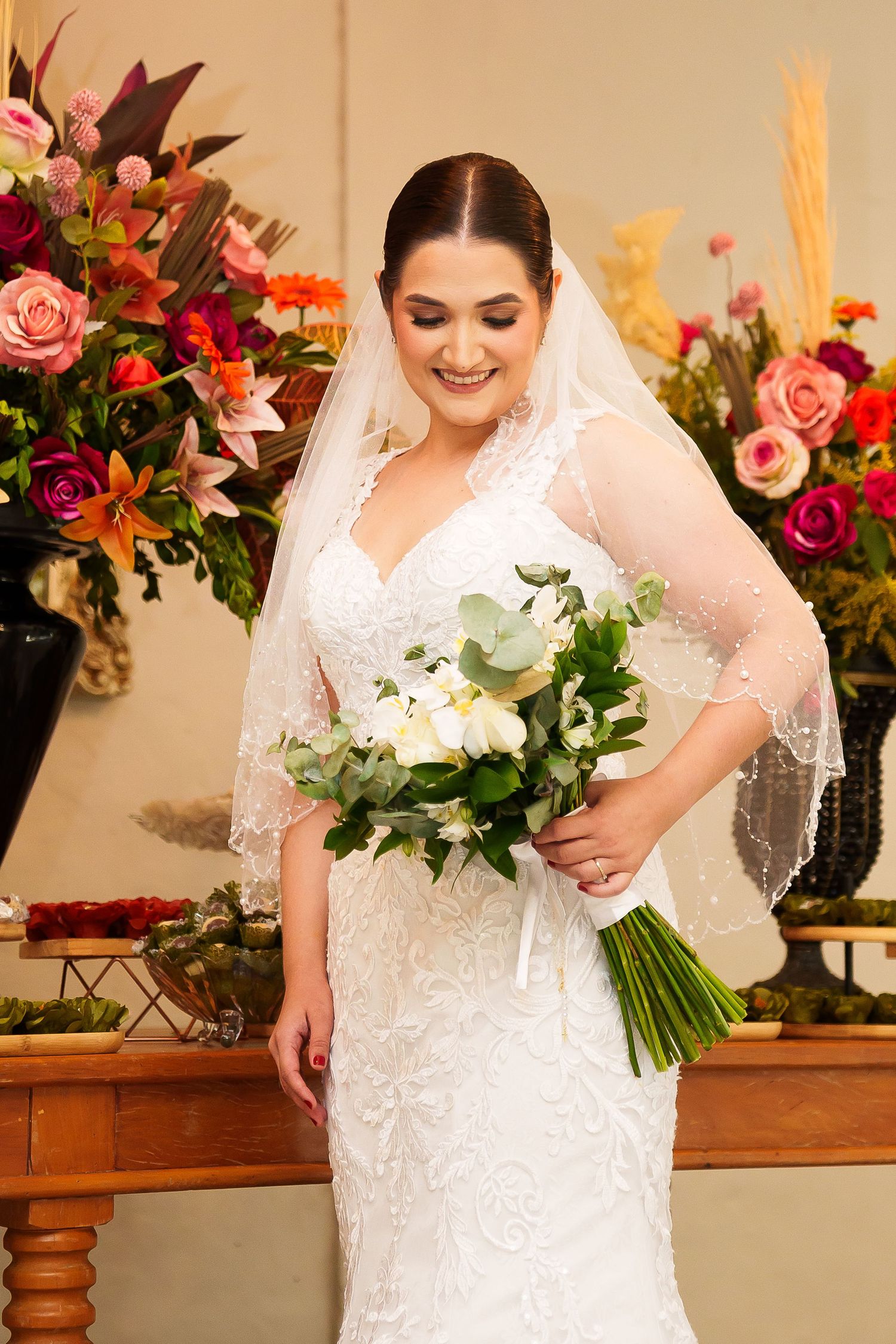 Smiling bride in lace wedding dress and veil holding white bouquet, standing before colorful floral arrangements.