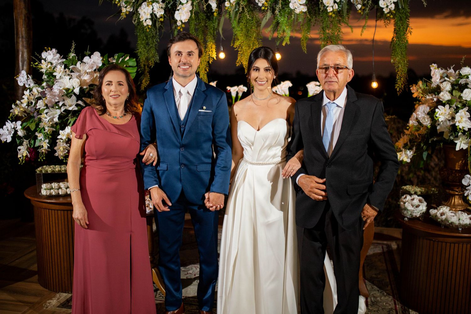 Wedding party of four posing together, bride in white gown, groom in blue suit, flanked by elegant older couple.