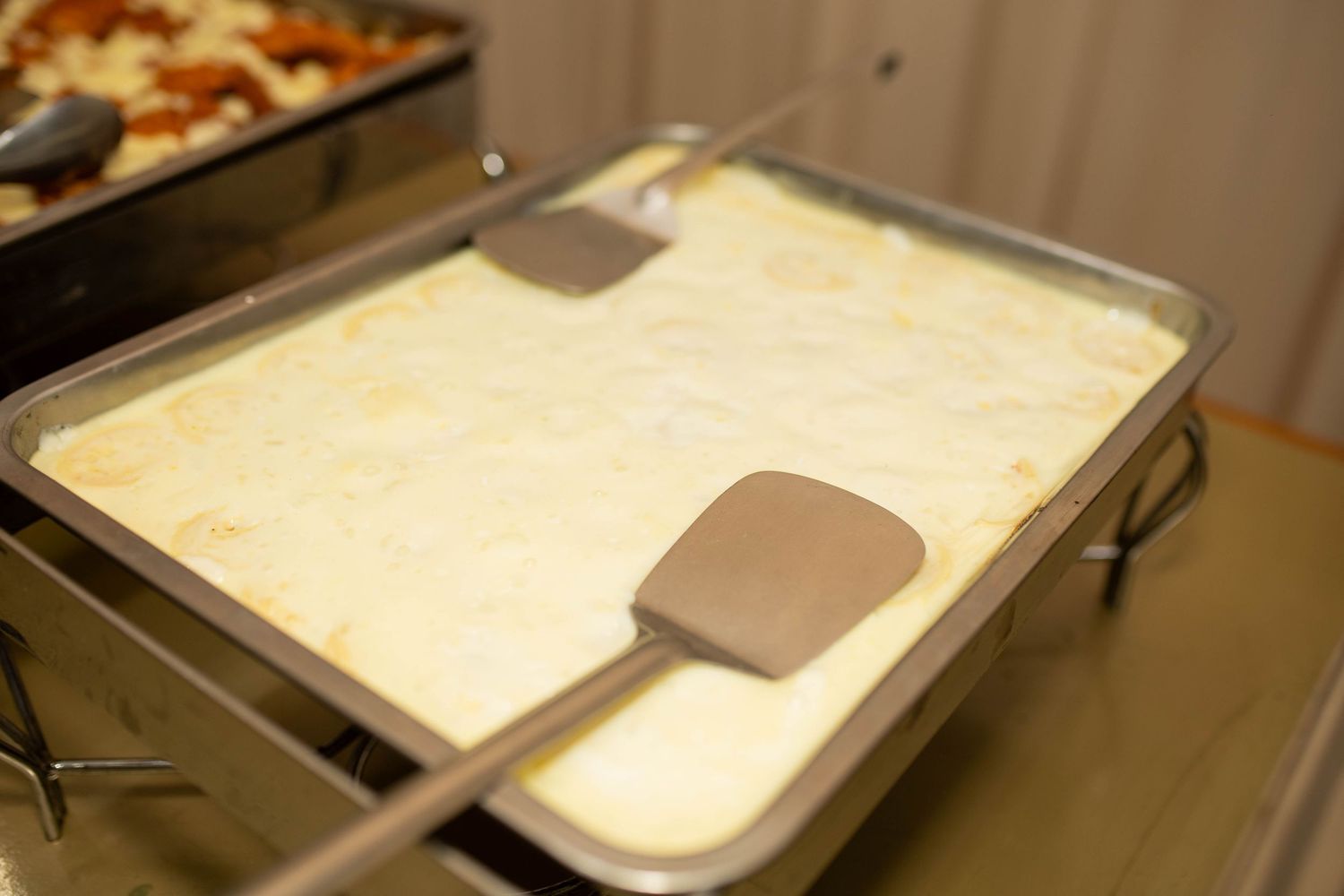 A large metal chafing dish filled with creamy lasagna or pasta bake, served with two spatulas at a buffet.
