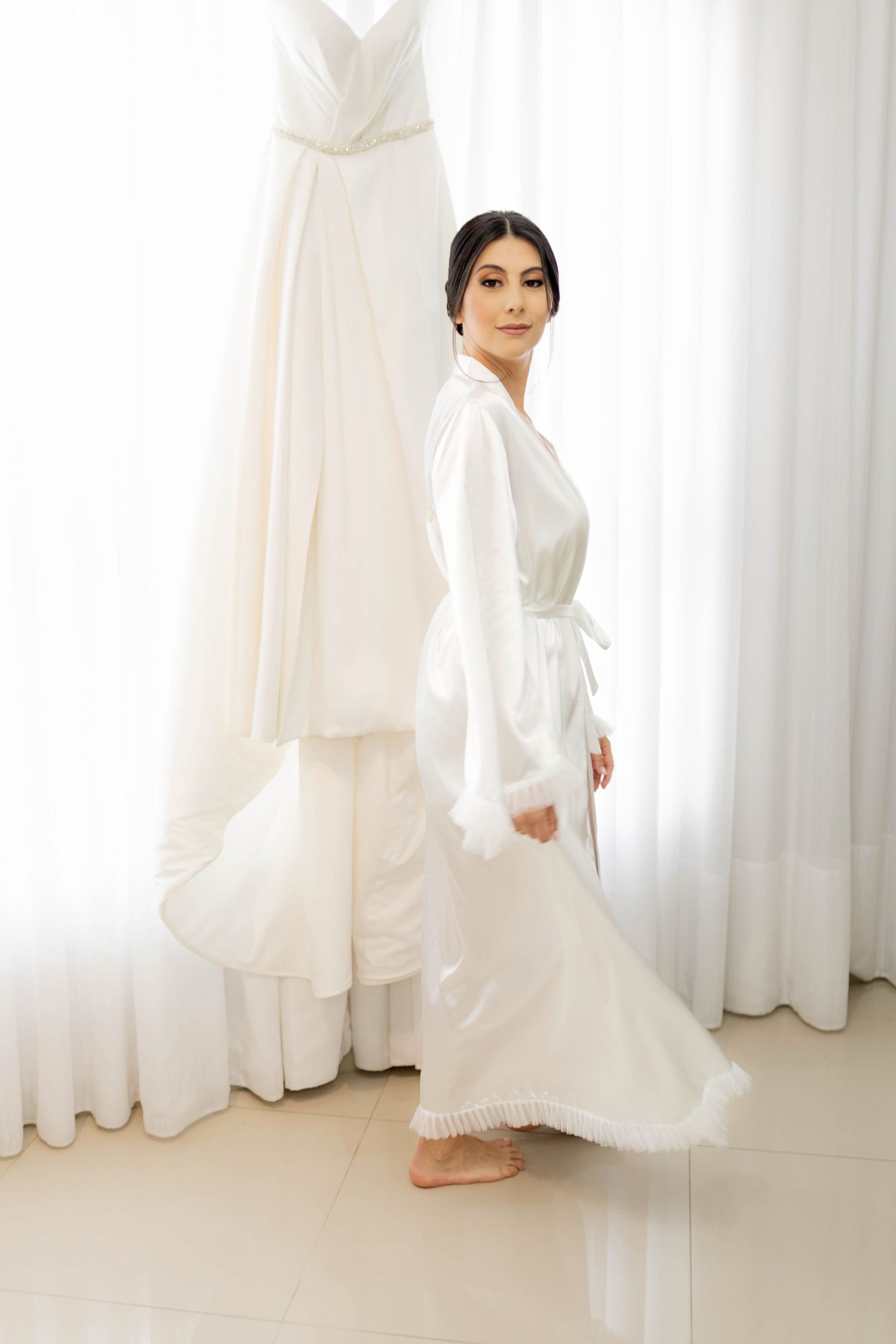 Bride in white satin robe with feather trim stands before her wedding gown hanging by sheer curtains.