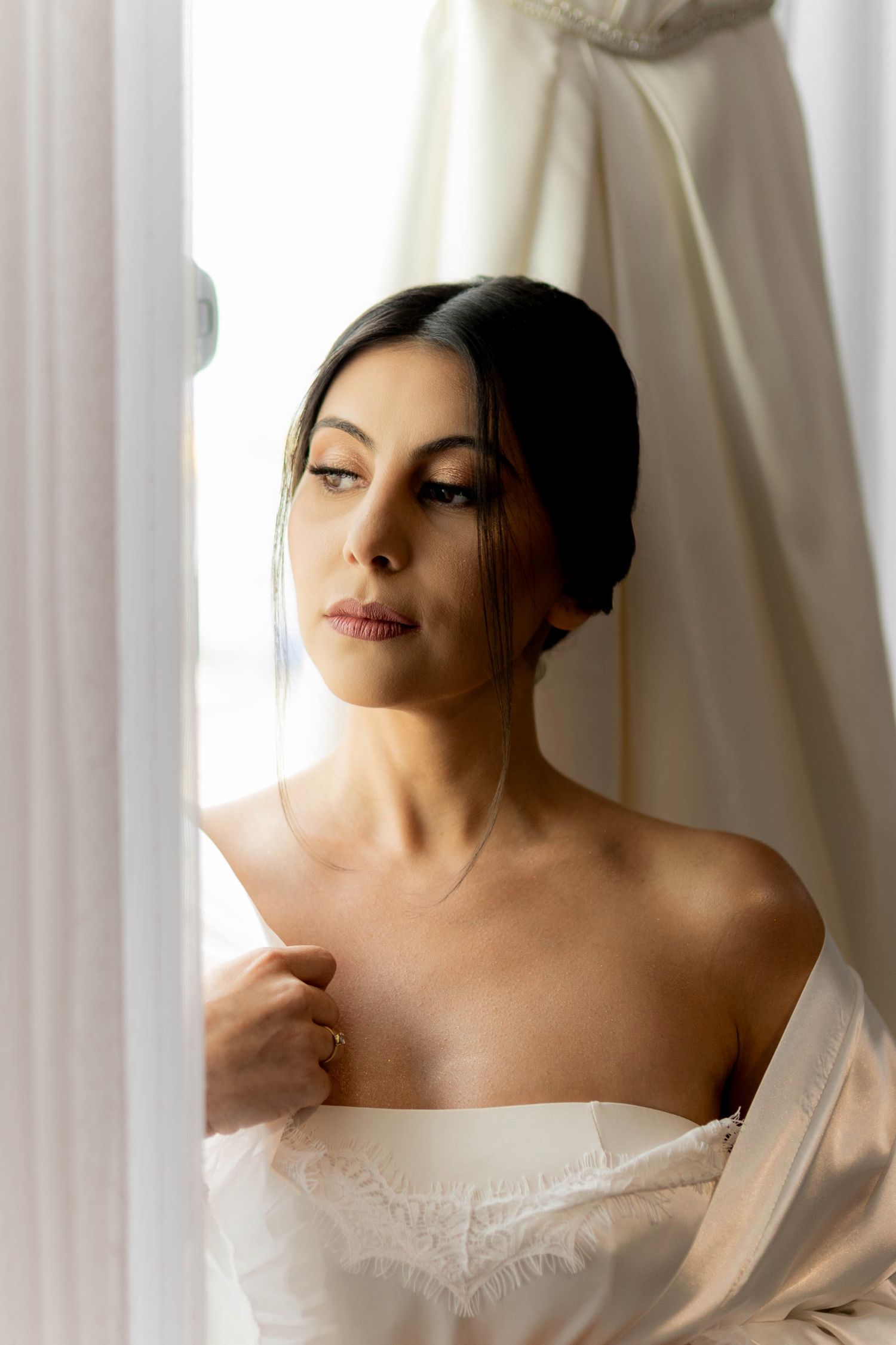 Elegant bride in white strapless gown gazing thoughtfully by a sunlit window with soft curtains.
