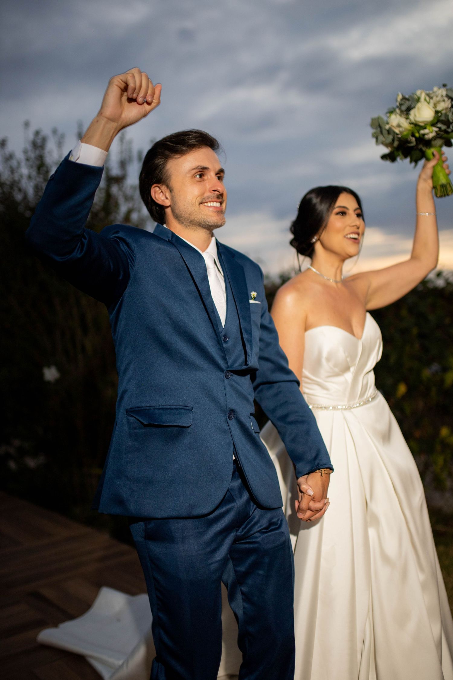 Joyful newlyweds celebrate outdoors, groom in blue suit raises fist, bride in white gown holds bouquet aloft.