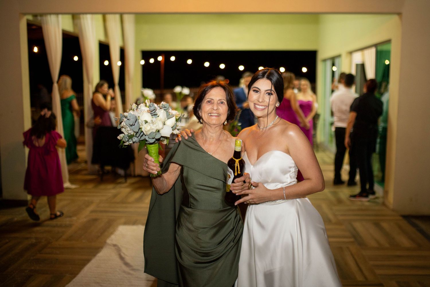 Bride in white strapless gown poses with older woman in olive dress holding bouquet at elegant wedding reception.