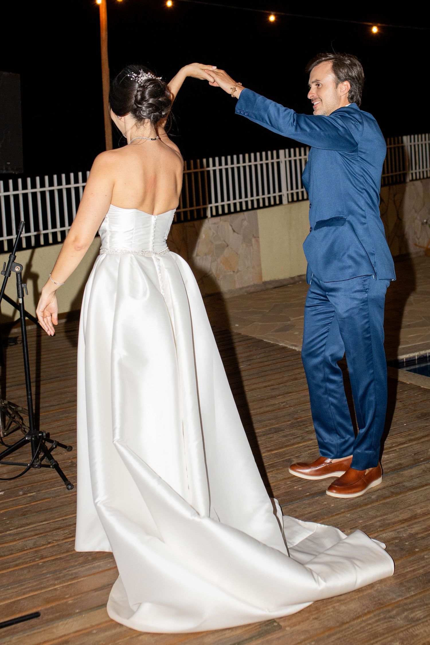 Bride and groom share their first dance outdoors at night, groom in blue suit spinning bride in white strapless gown.